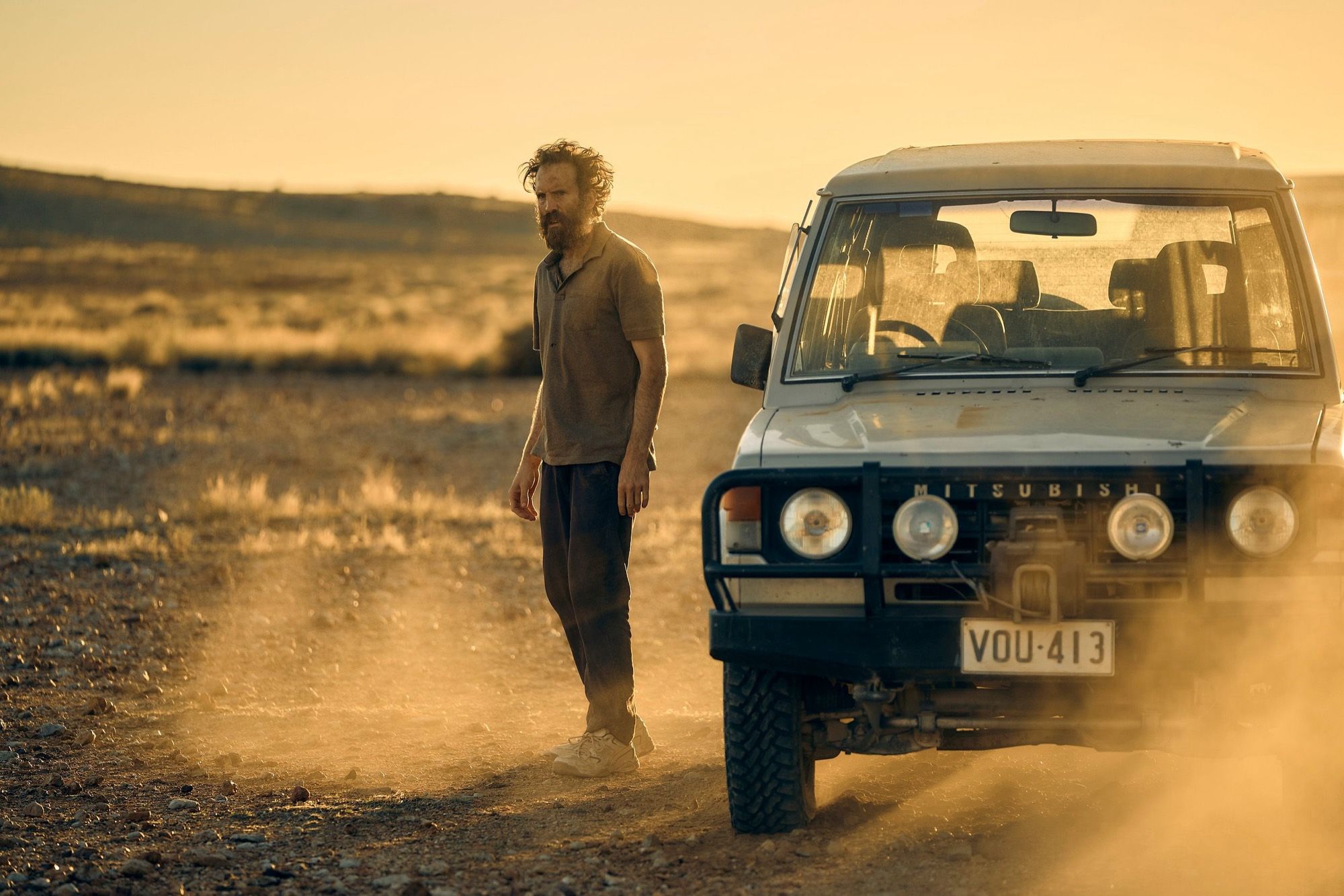 A bearded man stands beside an old four-wheel drive in a cloud of dust.