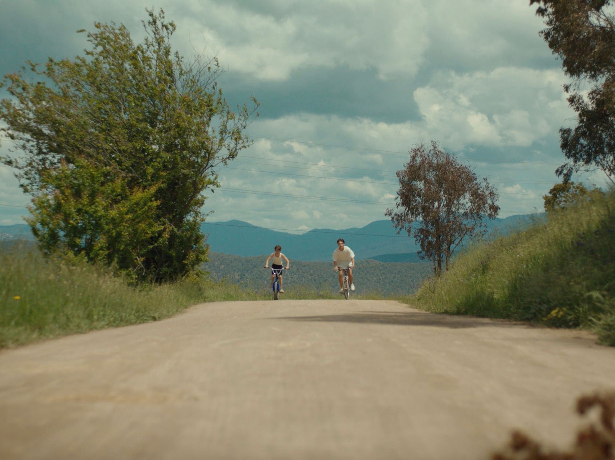 Two boys cycle down a dirt road through summer countryside.