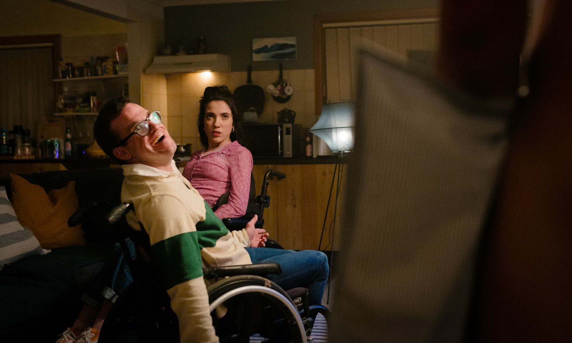 A young man in a wheelchair laughs in a dim living room; a young woman in a wheelchair looks on.