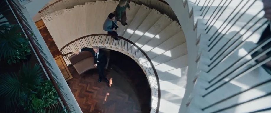 Seen from above, Sam Neill waits at the foot of a spiral staircase as two figures come down.