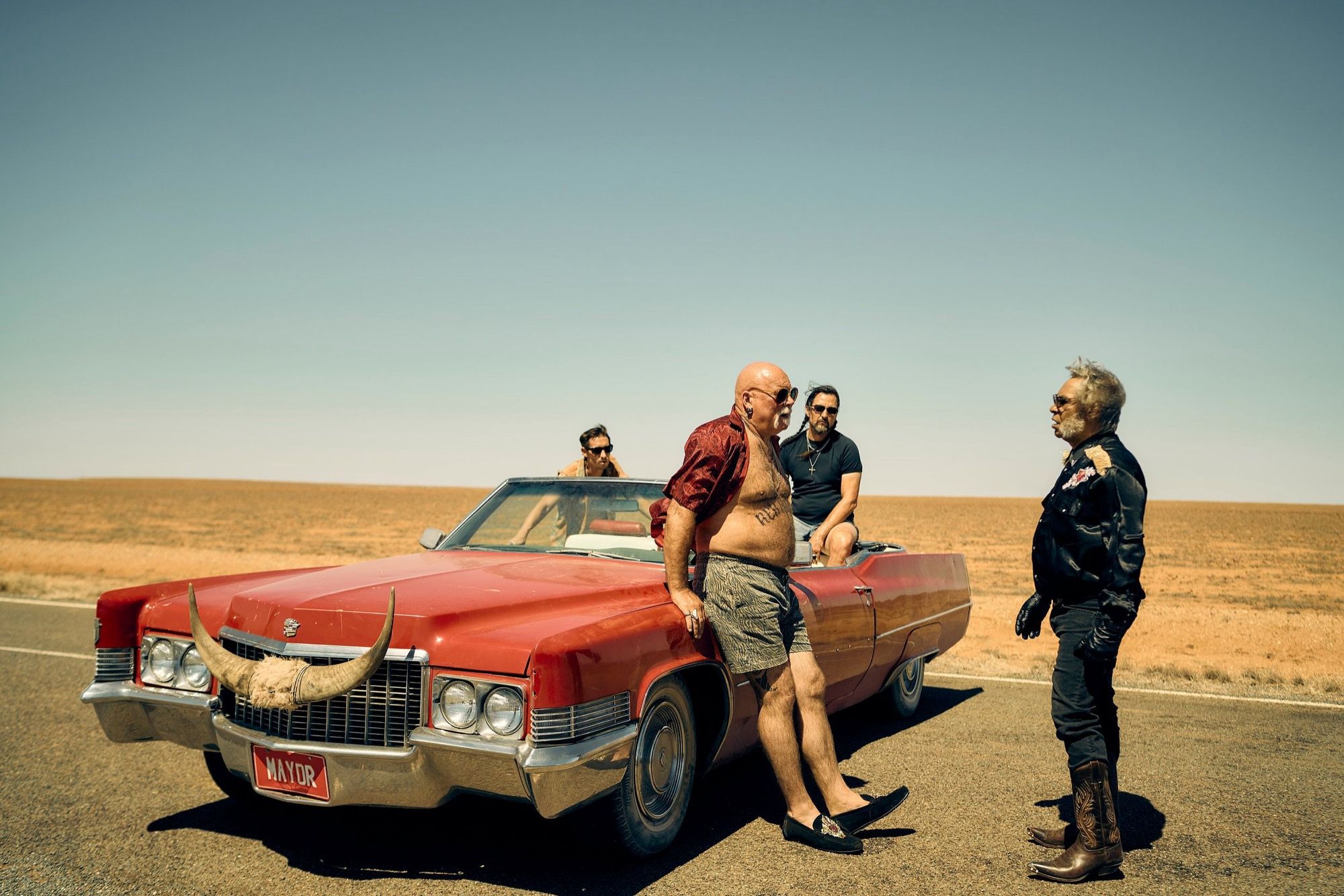 A shirtless man and three others around a red Cadillac with bull horns on its bonnet, on a desert highway.