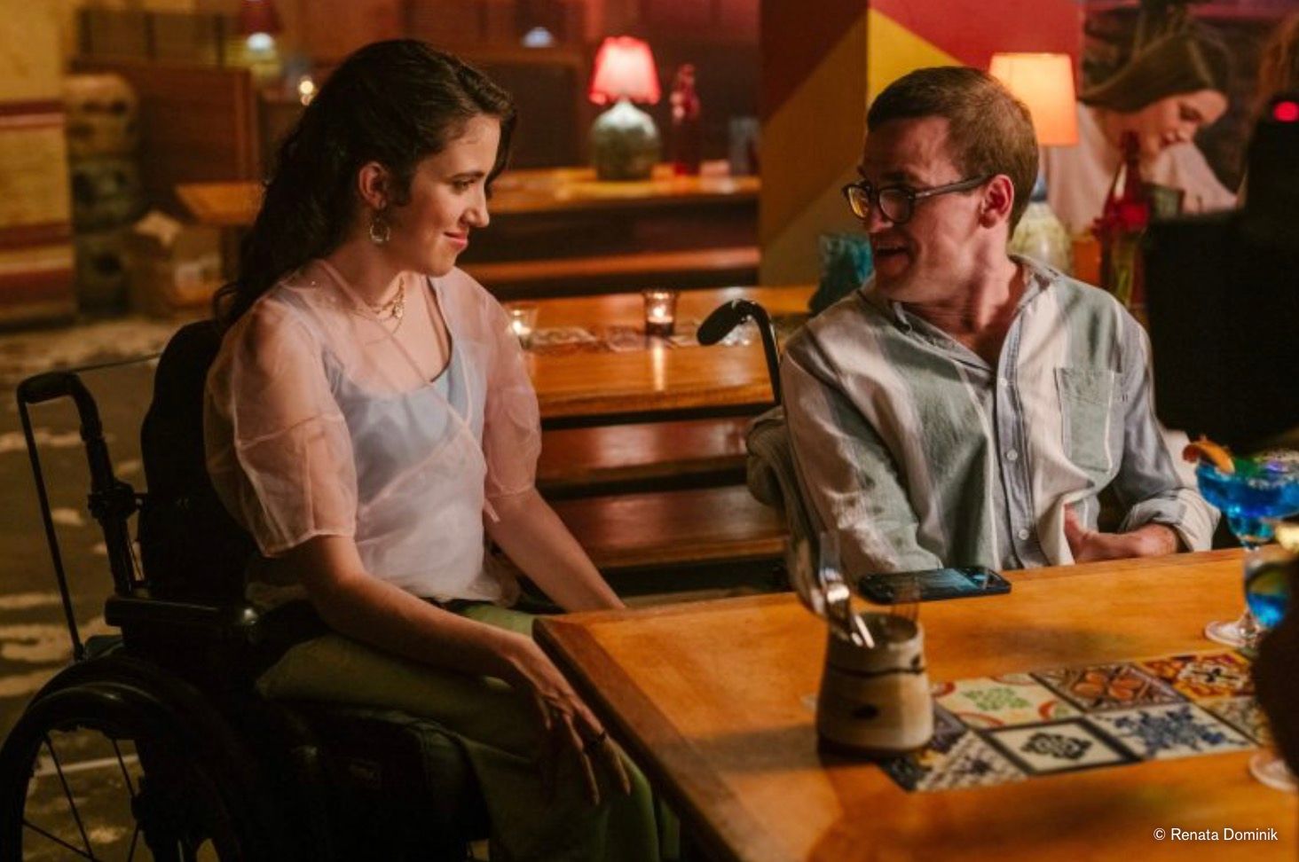 The two leads talk across a table in a tiled bar.