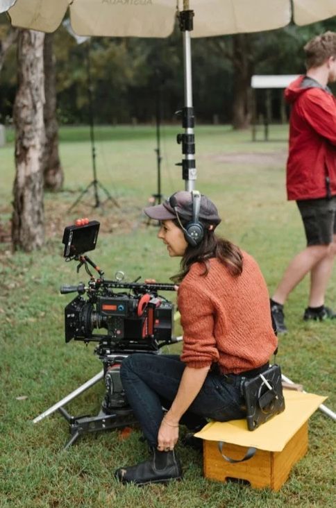 Behind the scenes: a woman in a cap and headphones sits beside the camera on a park location.