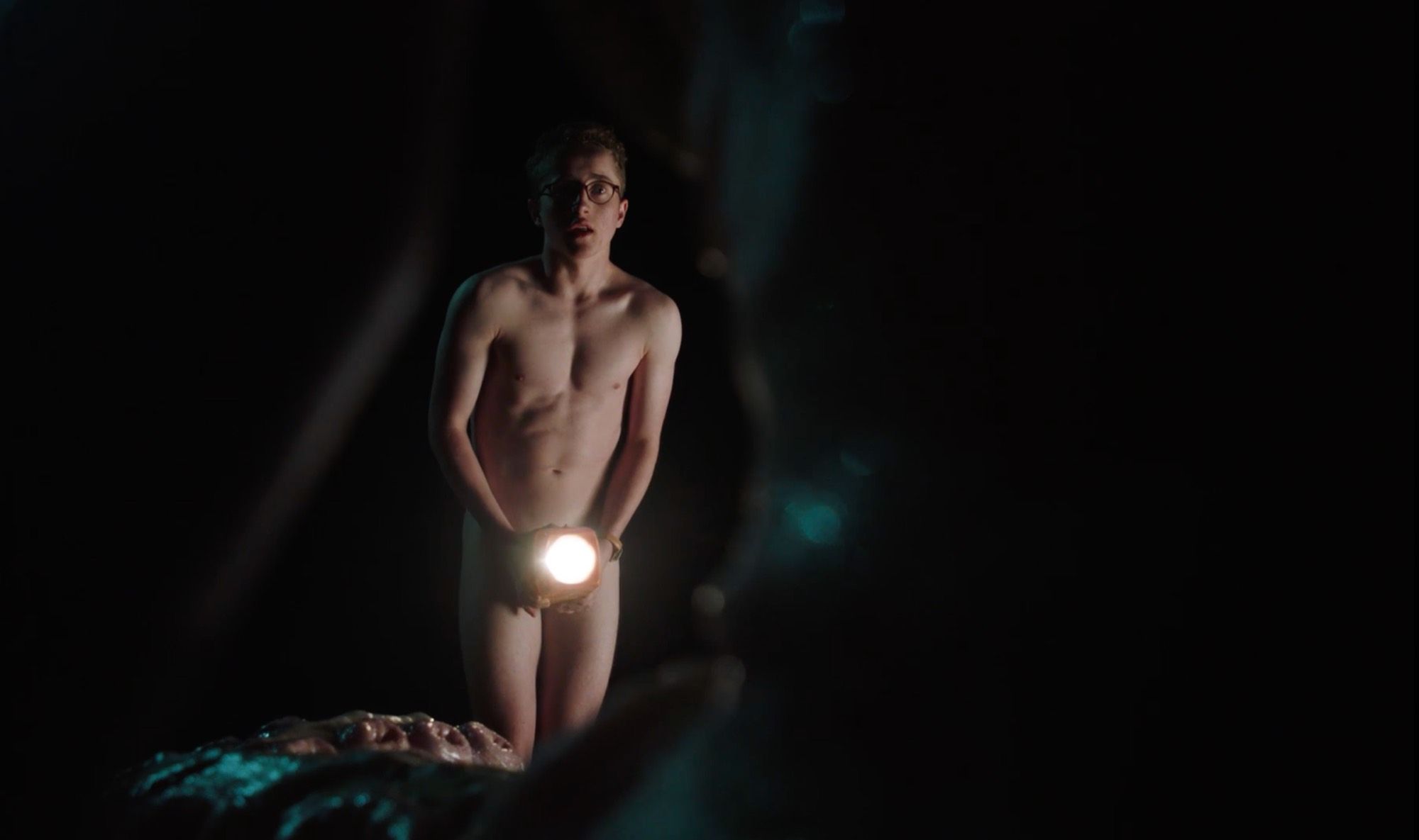 A naked young man in glasses, lit by a torch in the dark.
