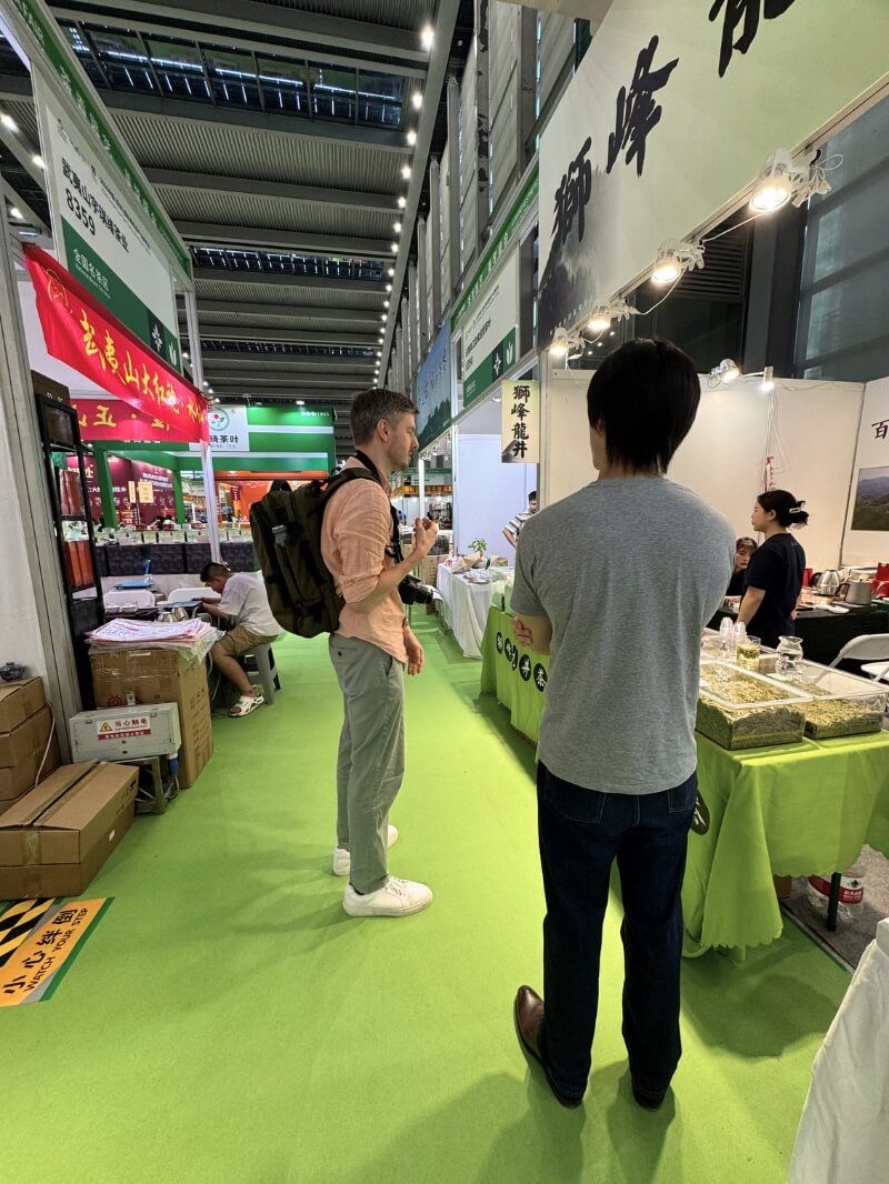 Two men stand talking in an aisle at the tea expo, a green-carpeted walkway between booths, trays of green tea on a table beside them.
