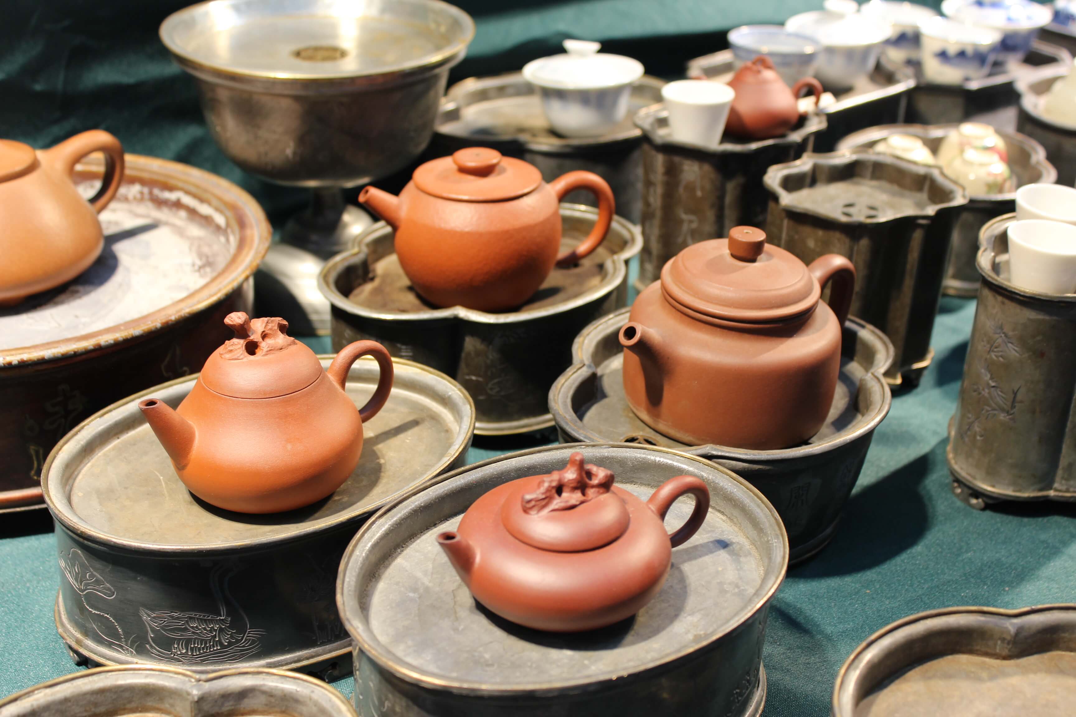 Small clay teapots on pewter saucers arranged on a teal cloth, with cups and larger vessels behind.