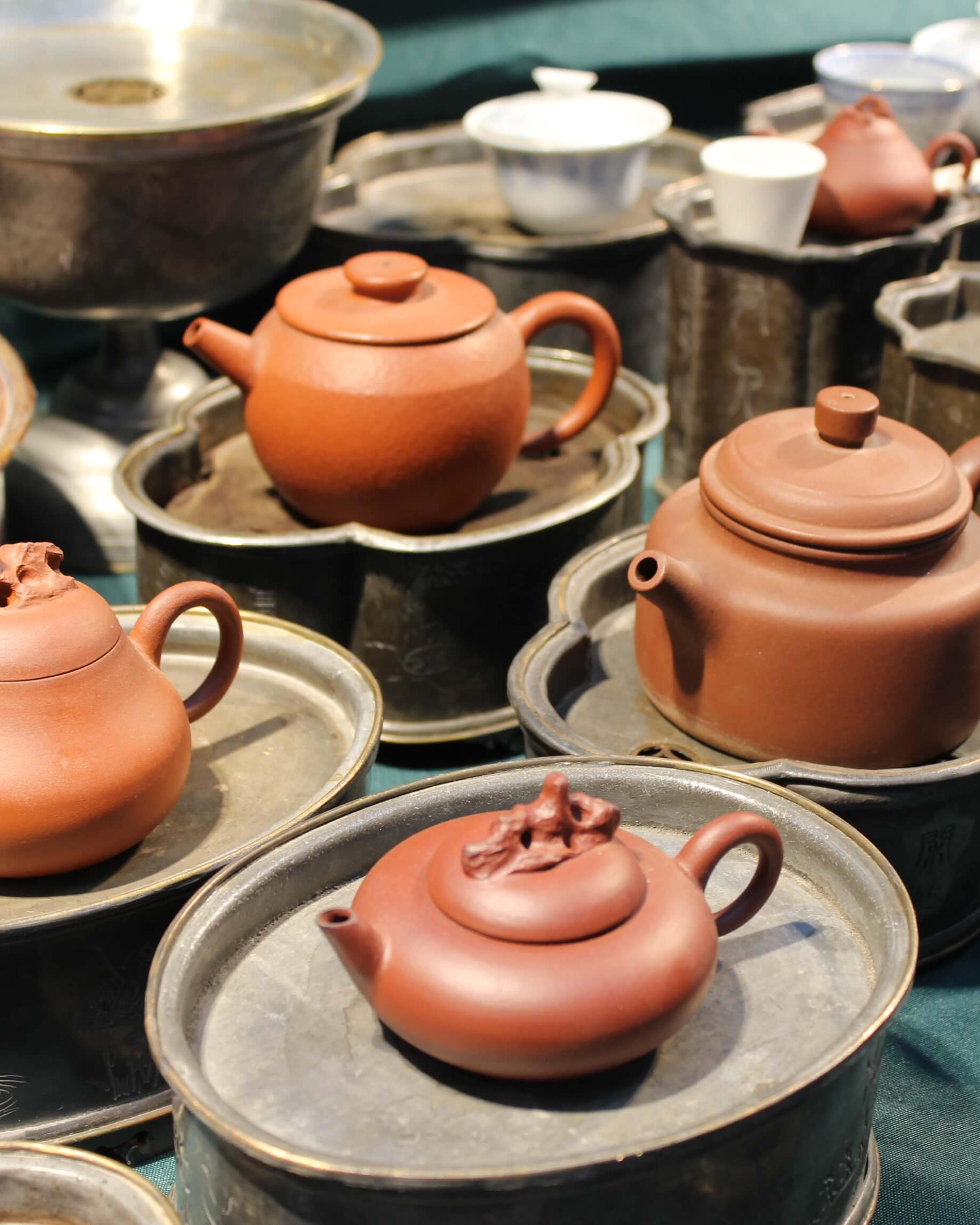 Small clay teapots on pewter saucers arranged on a teal cloth, with cups and larger vessels behind.