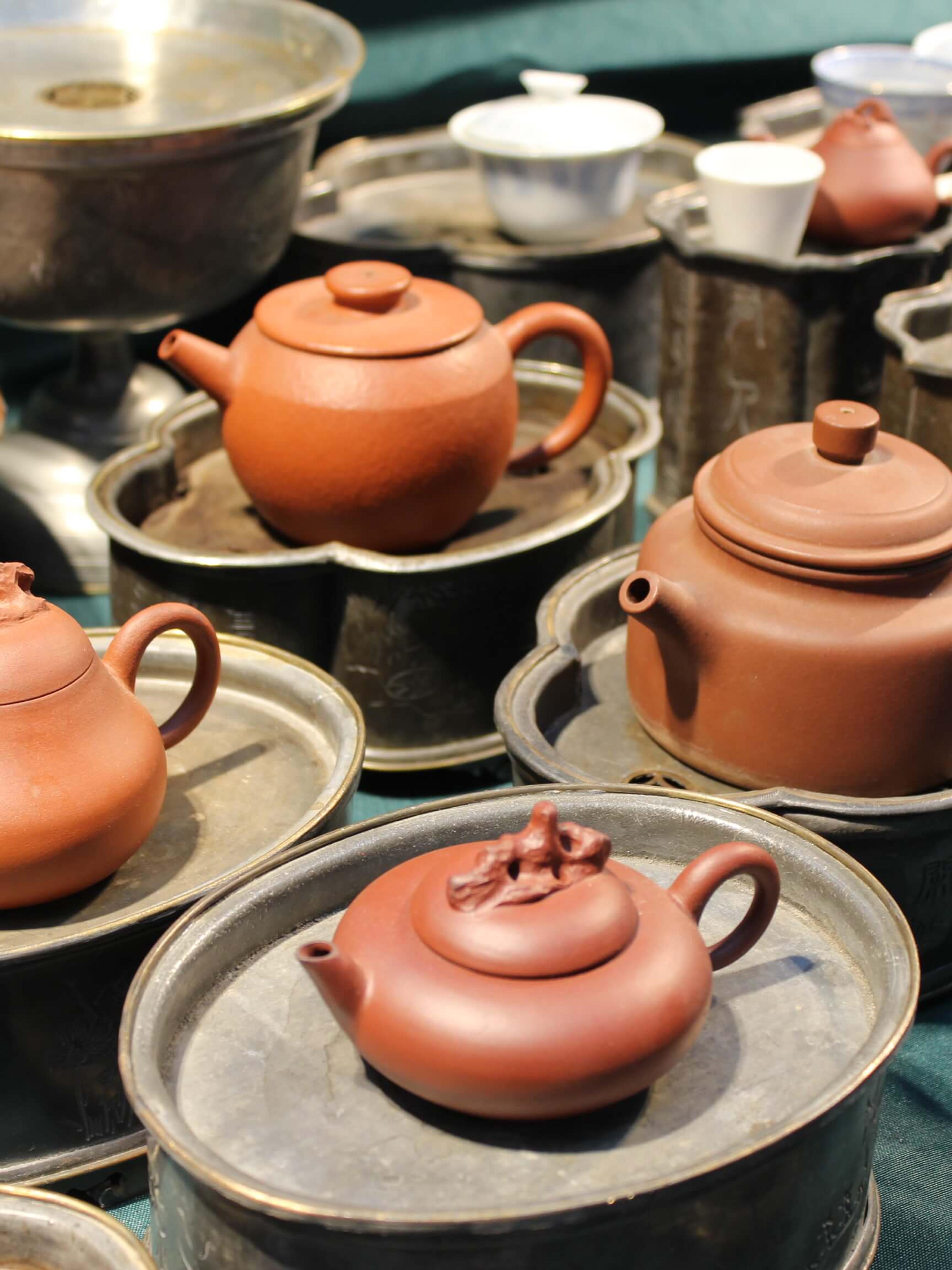 Small clay teapots on pewter saucers arranged on a teal cloth, with cups and larger vessels behind.