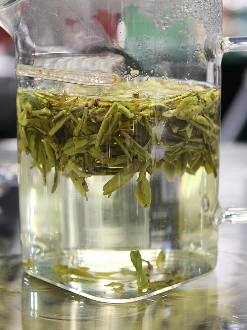 A glass pitcher of pale green tea, leaves steeping above clear liquor.