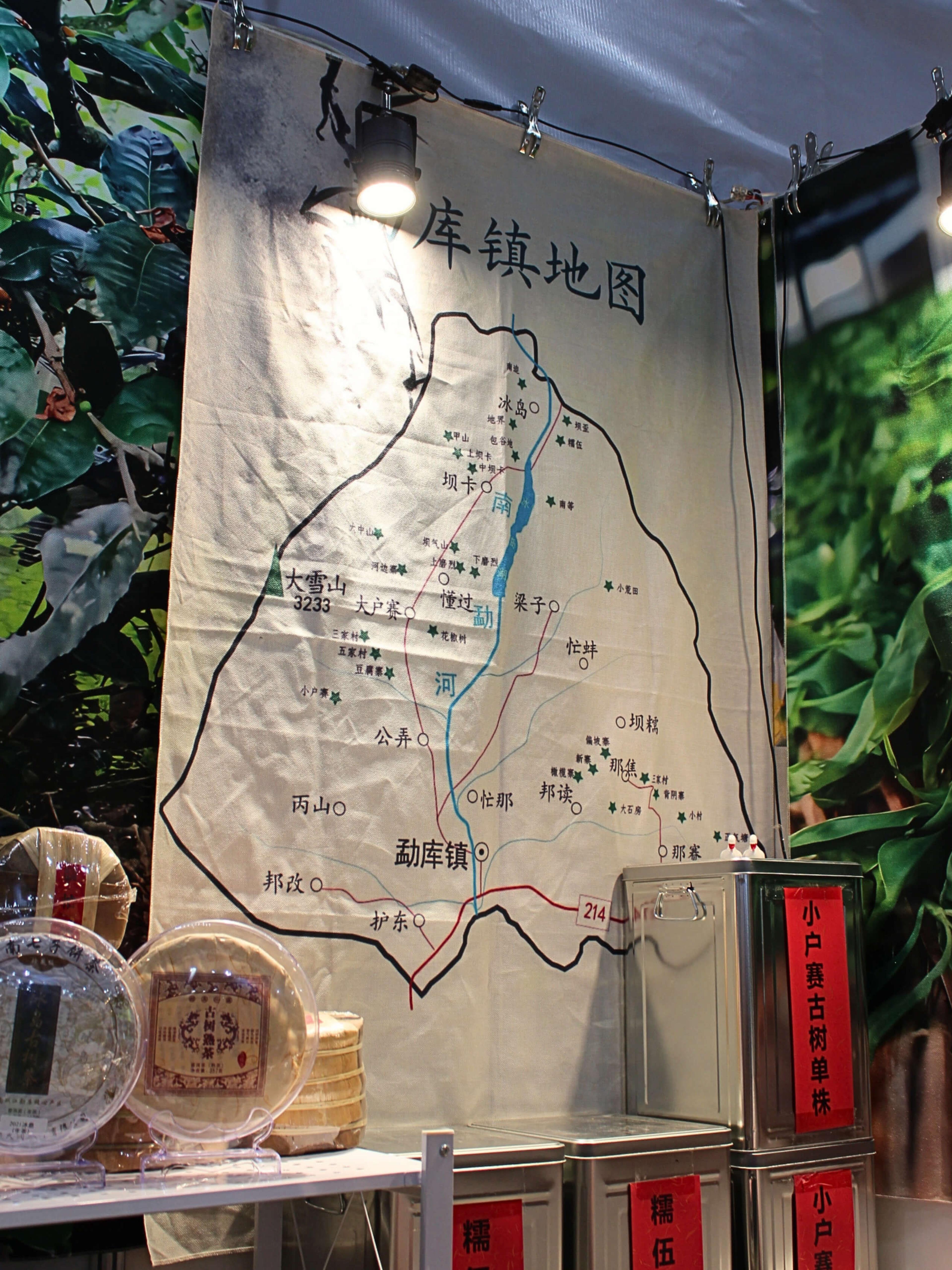 A map of Mengku town in Lincang, Yunnan, hanging at the back of Fu Yuanyuan’s stand. Villages are marked along the Nanmeng river valley, with Mengku town at the southern end and Bingdao at the northern end. Tins of tea sit on shelves beneath it.