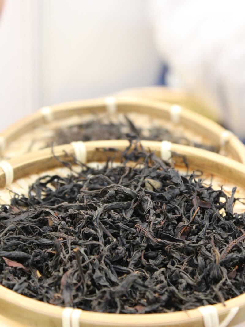 A shallow bamboo basket holding dark, tightly rolled tea leaves, set on a table beside a glass.