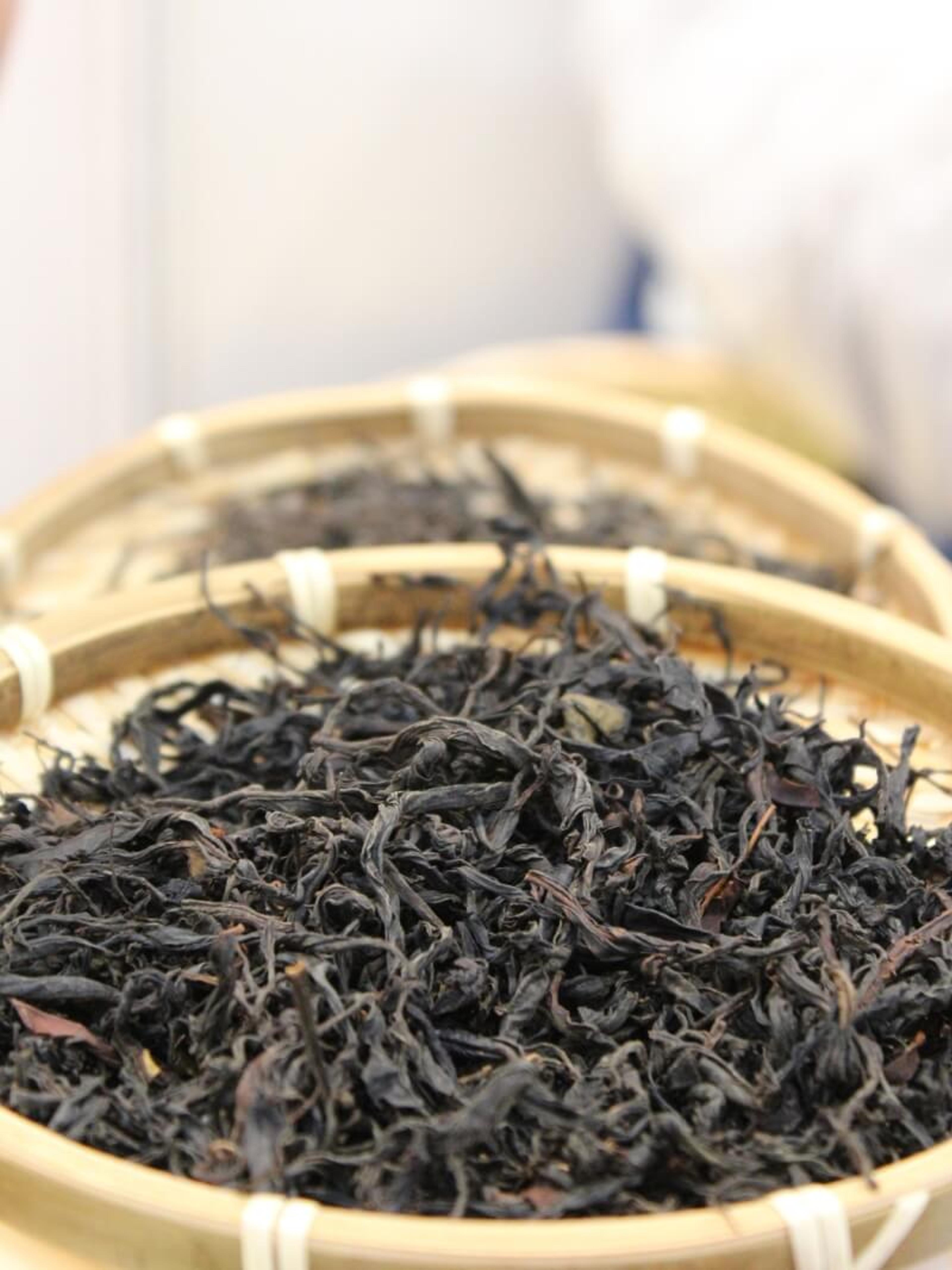 A shallow bamboo basket holding dark, tightly rolled tea leaves, set on a table beside a glass.