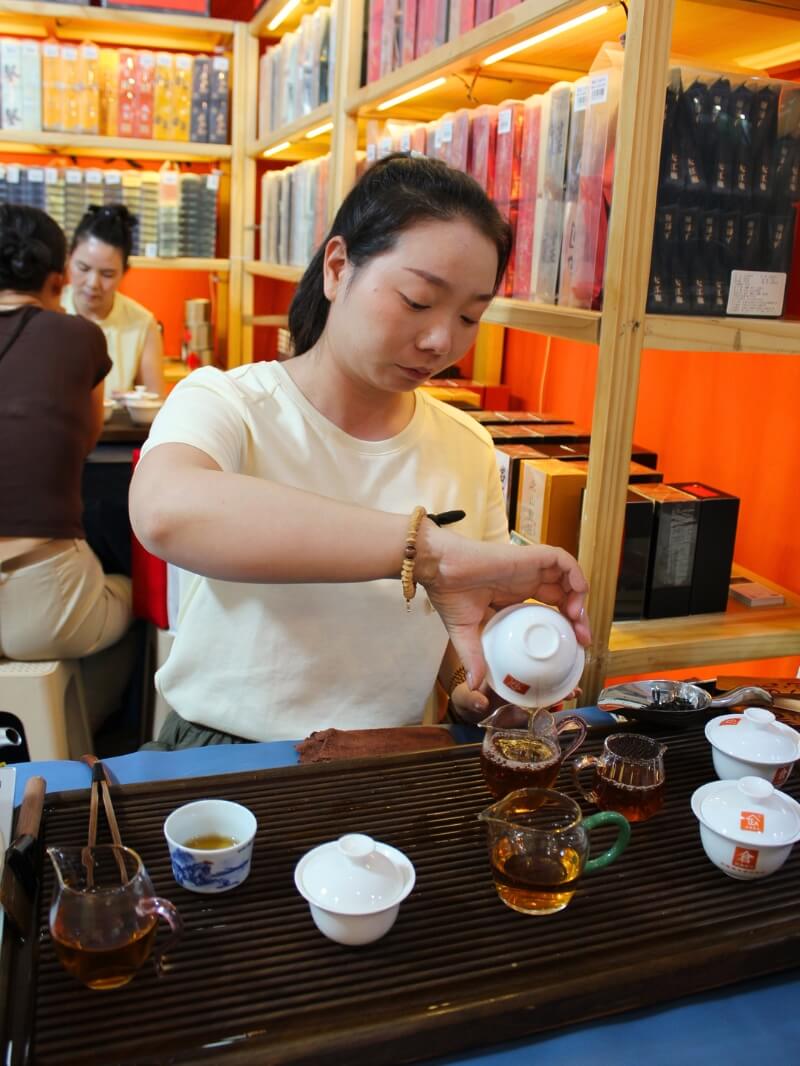 A woman pours tea from a gaiwan at a tasting table, shelves of boxed rock oolong behind her.