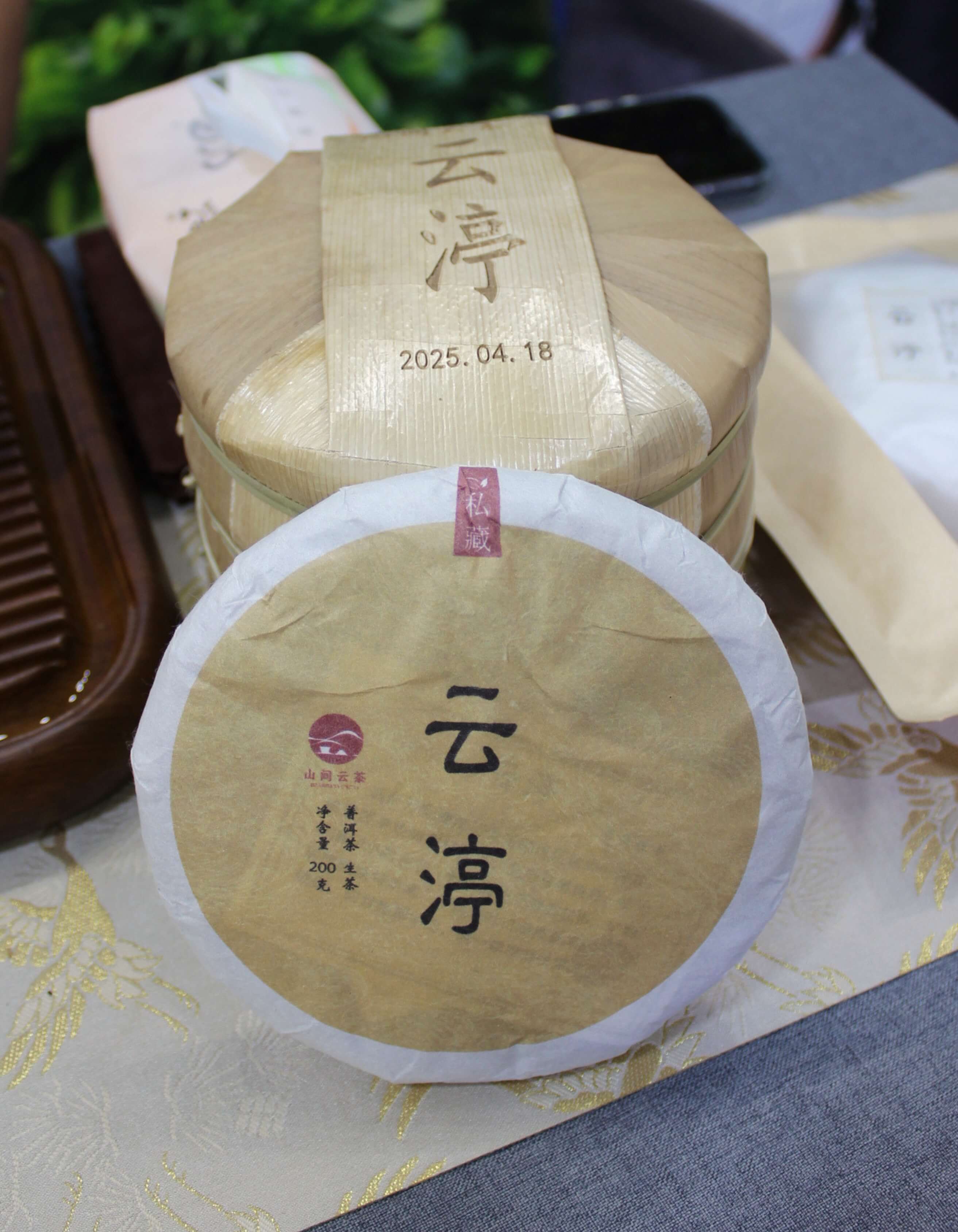 A round puer cake in cream paper wrapping, stamped with a date, on a patterned cloth.