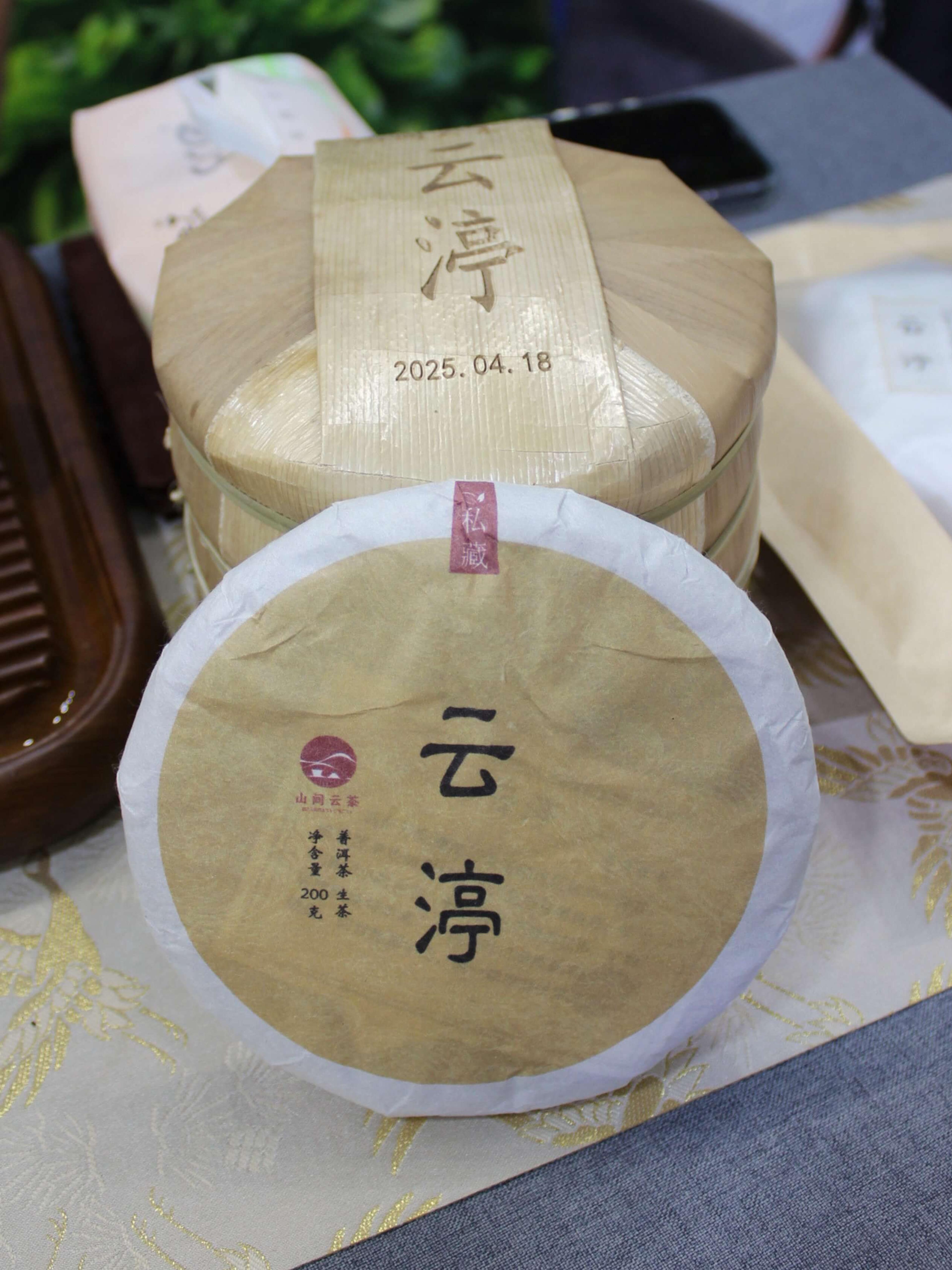 A round puer cake in cream paper wrapping, stamped with a date, on a patterned cloth.