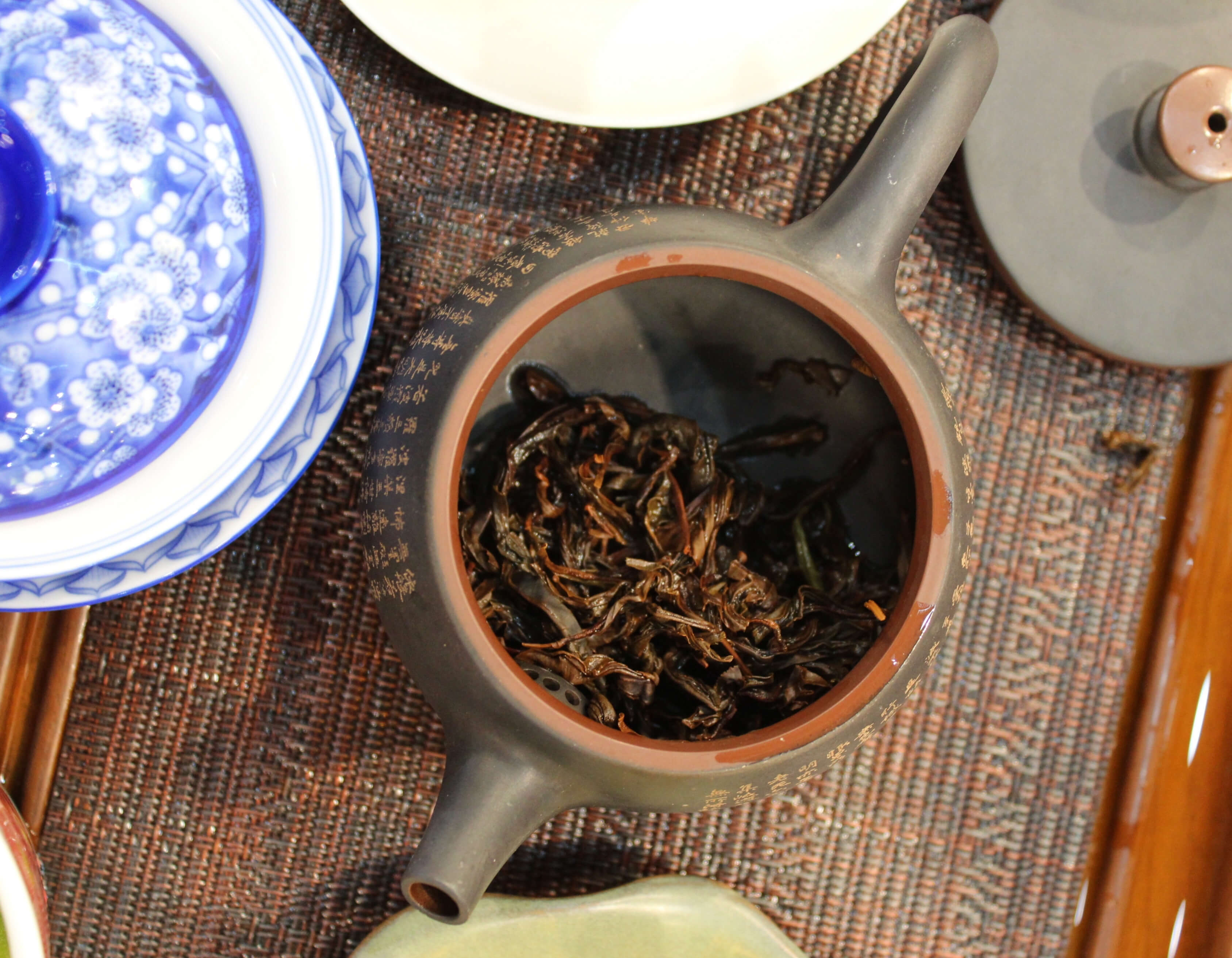 Overhead view of a black clay teapot with its lid off, gold characters around the body and a terracotta rim, spent tea leaves inside.