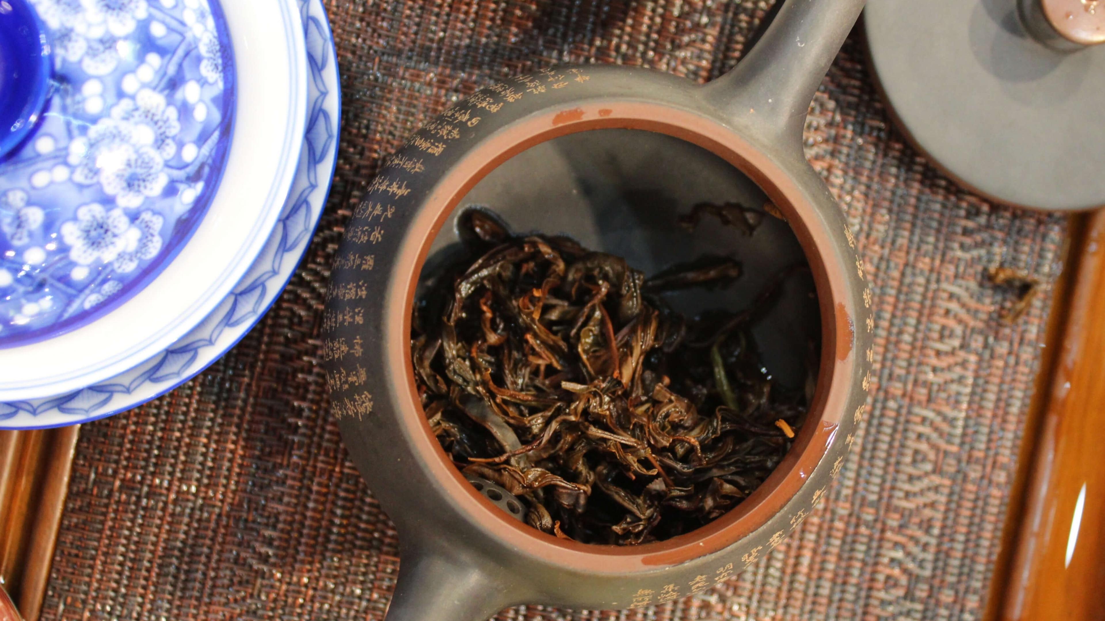 Overhead view of a black clay teapot with its lid off, gold characters around the body and a terracotta rim, spent tea leaves inside.