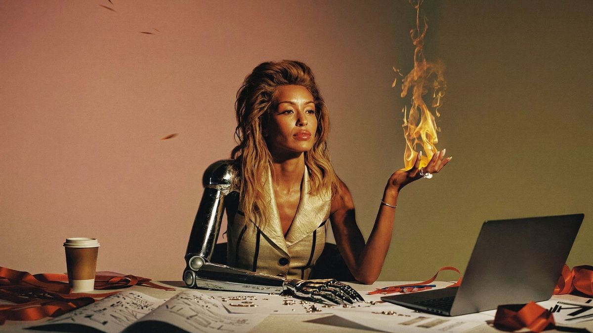A woman at a cluttered design desk holds a flame of creative energy in her human hand, while her other arm is a chrome robotic arm resting on the work.