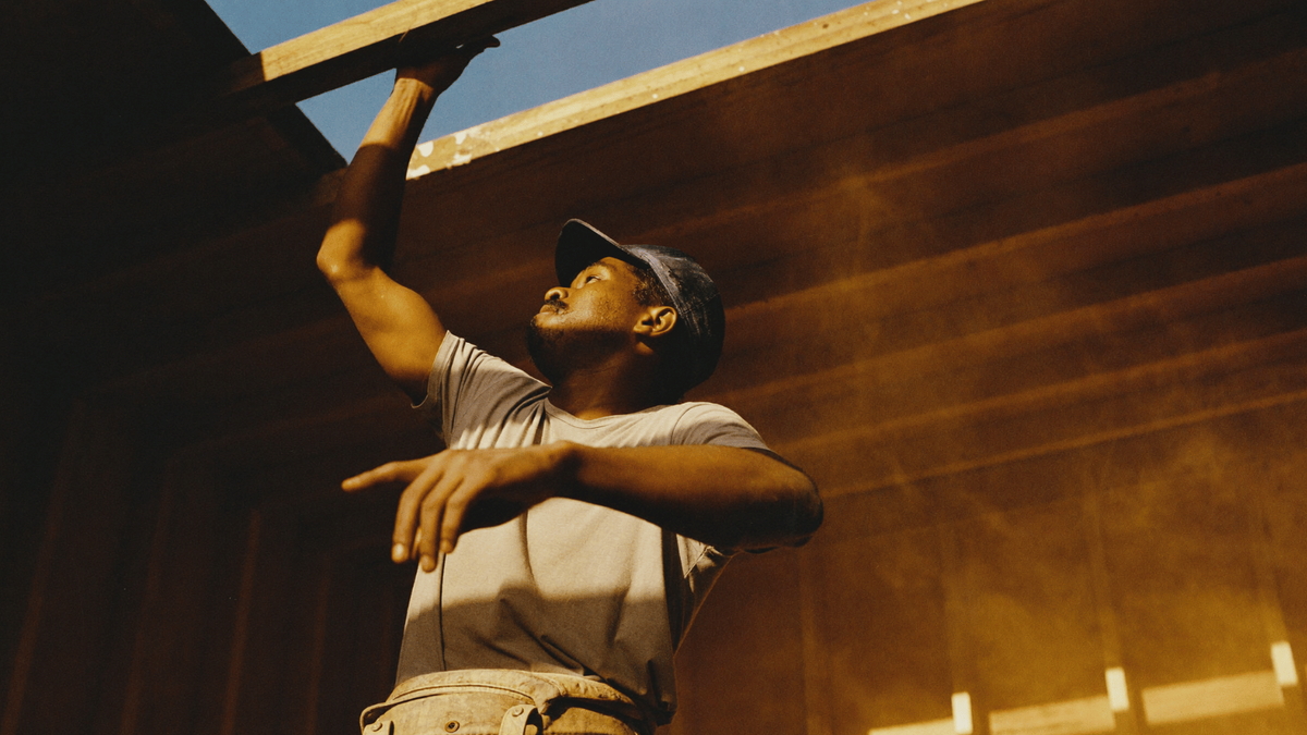 A contractor in a work cap reaching up to position a wooden beam during framing, lit by warm afternoon sun.