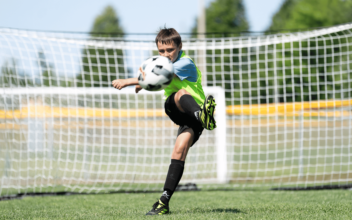 A young boy in soccer gear kicking a ball toward the camera on a field, with the goal net behind him.