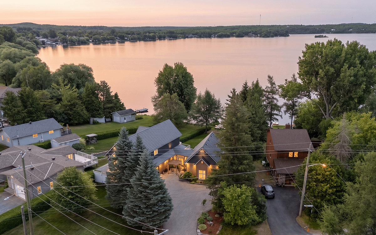 Aerial drone photo of a lakefront property at sunset, showing the home, driveway, and surrounding neighbourhood along the shoreline.
