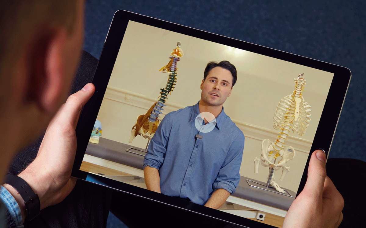 Tablet held by a person, displaying a video of an osteopathy practitioner speaking to camera in a clinic with anatomical spine and skeleton models in the background.