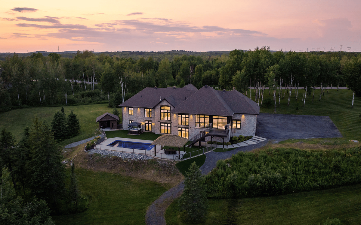 Aerial drone photo of a large stone luxury home with a lit interior, pool, and gazebo at sunset, surrounded by forest and open lawn.