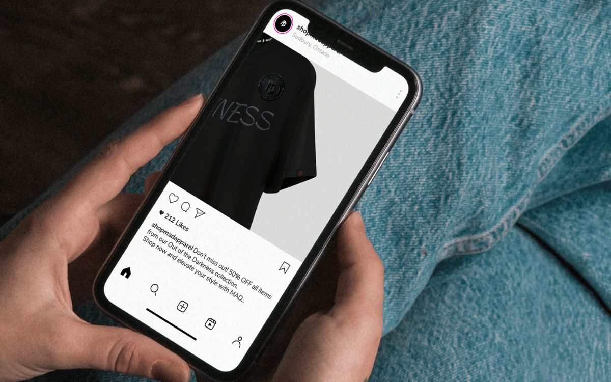 Hand holding a smartphone displaying an Instagram shop ad for a black apparel brand, showing a product photo and a 50% off promotion caption.