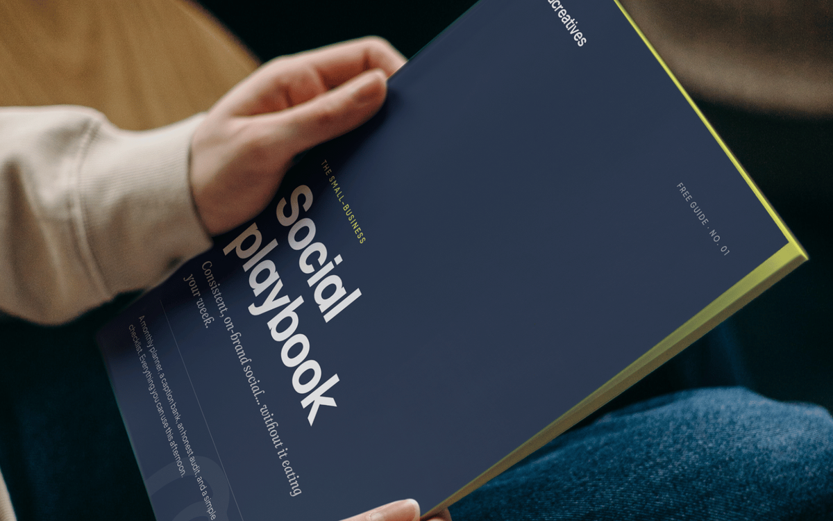 A photo mockup of a hand holding "The Small-Business Social Playbook" guide, a navy cover with yellow-green edges reading "Consistent, on-brand social... without it eating your week."