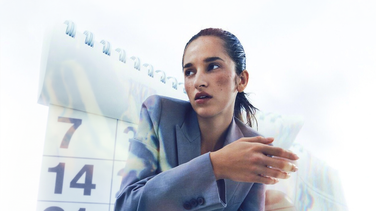 A woman in a grey blazer looks over her shoulder in front of an oversized wall calendar showing the 7th, 14th, and 21st.