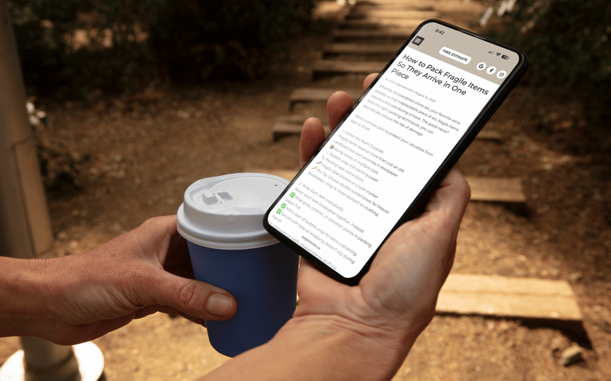A phone showing a We Got Moves blog post on packing fragile items, held next to a coffee cup outdoors.