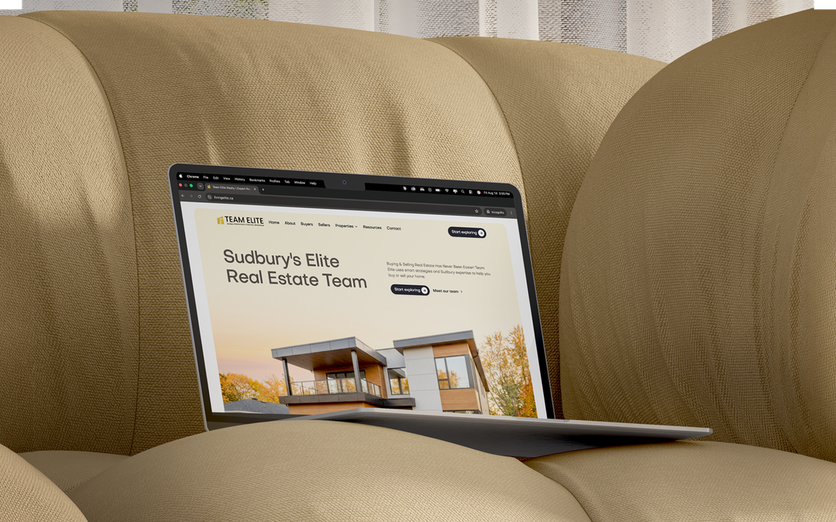 Laptop resting on a tan couch displaying the Team Elite Realty website homepage, featuring the headline 'Sudbury's Elite Real Estate Team' and a modern home exterior photo