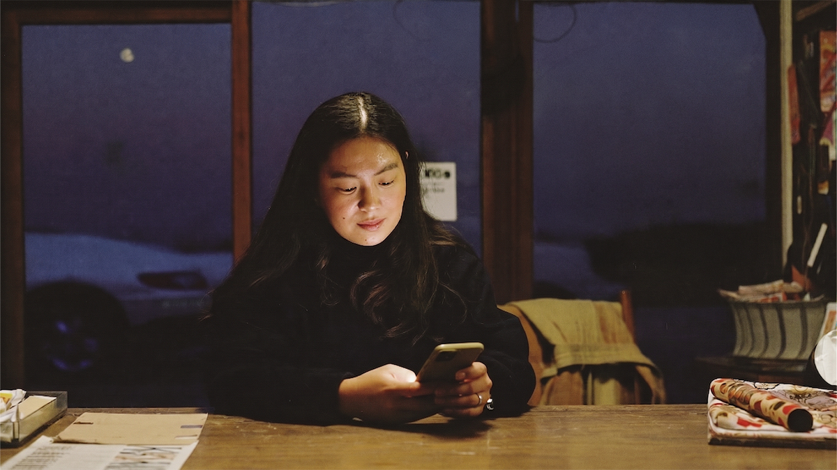 A shop owner looking at her phone behind a wooden counter, blue evening light in the windows behind her.