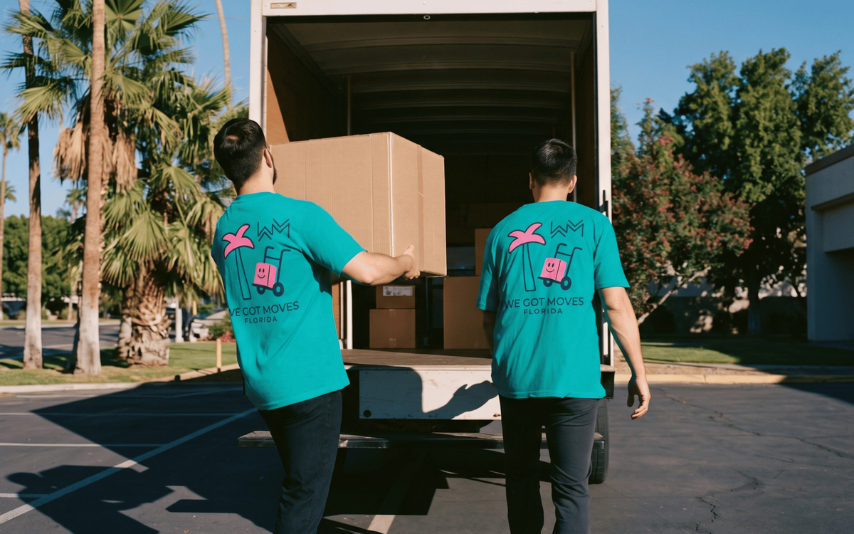 Two We Got Moves Florida movers in teal branded shirts carrying a box into a moving truck, palm trees in the background.