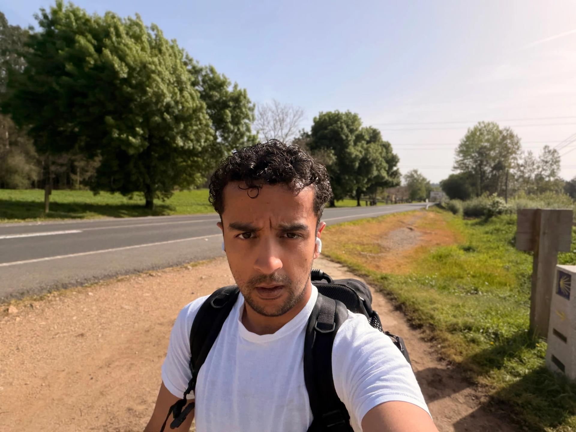 me, exerted, walking the Camino, on a dirt footpath with fields and woods and a road in view.