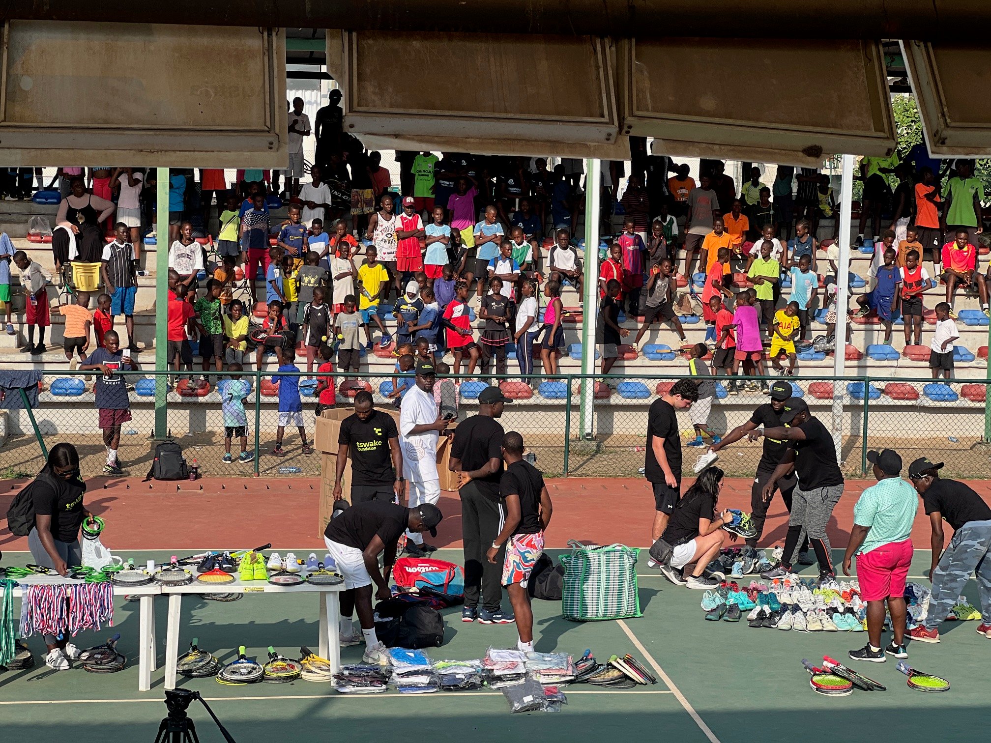 Kids fill the stadium stands while volunteers lay out rackets, shoes, and kits for distribution