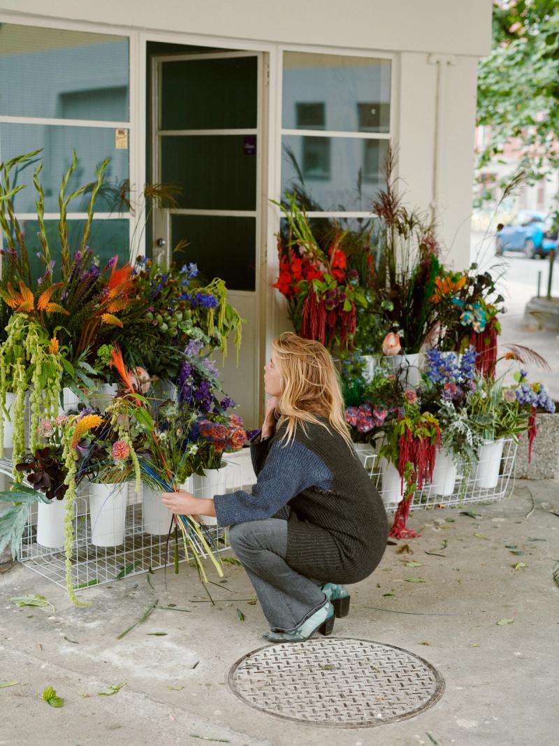 Karen Pederstad (Bukett!) crouches in front of a flower shop with flowers displayed outside.