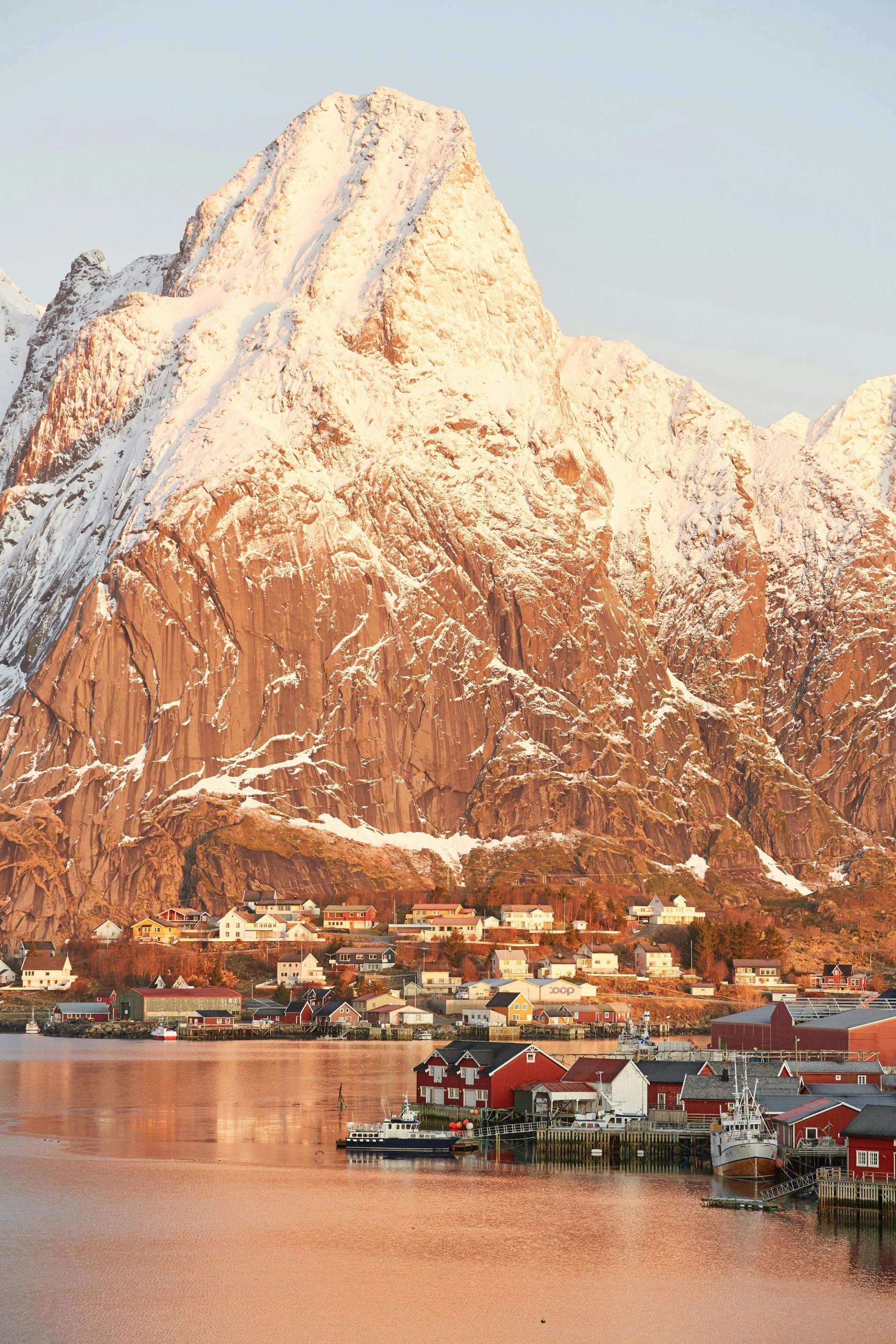 Snow-capped mountains rising above the red fishing cabins and still water of a Lofoten fjord at golden hour