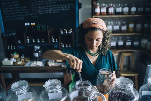 Herbalist scooping dried herbs from glass jars at her apothecary shop counter