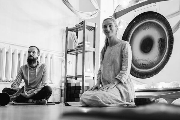 Black and white photo of two people seated cross-legged beside a large gong in a sound healing studio