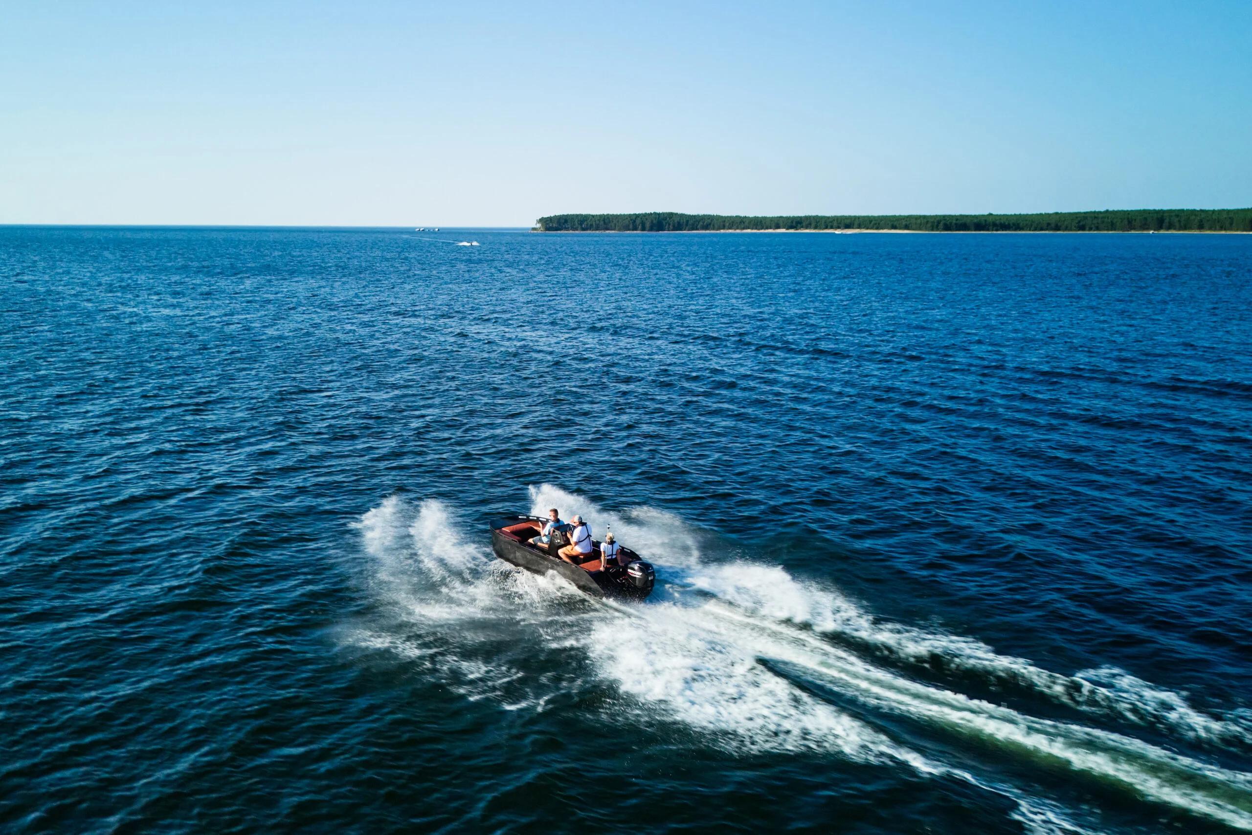 Aerial view of a black workboat planing across open blue sea toward a distant forested shore