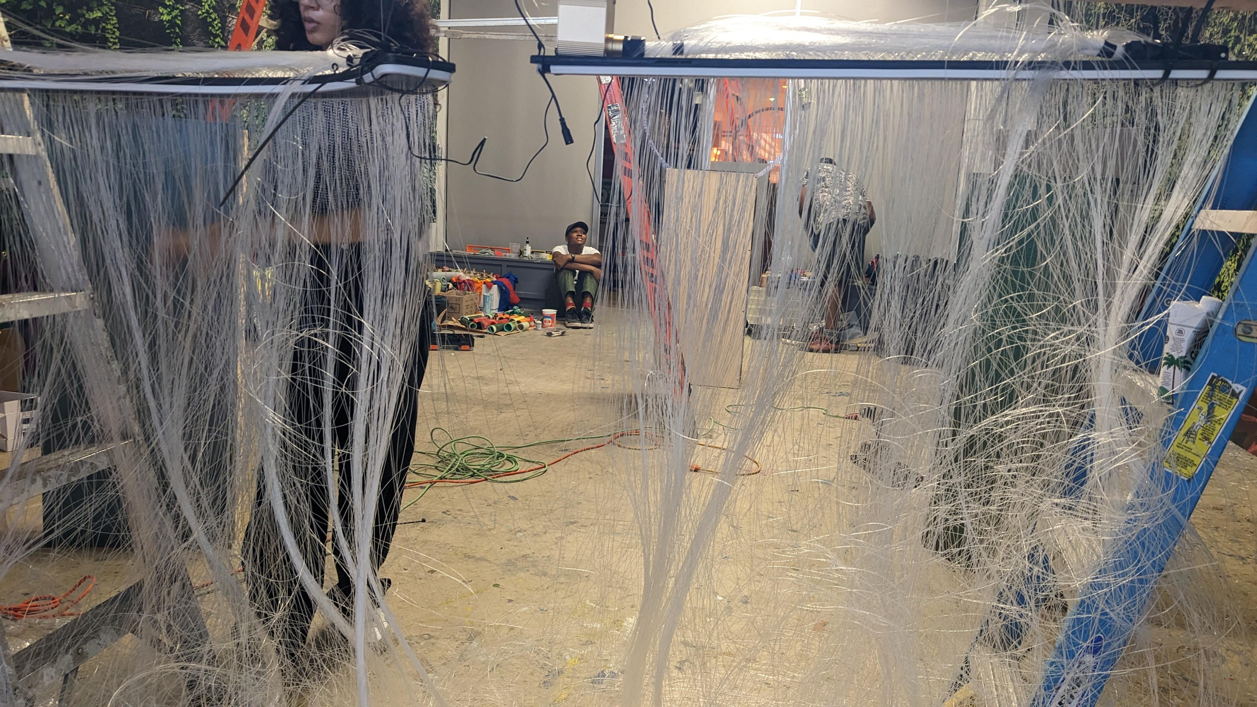 Photo by eo Studios. Photo of a gallery space during the installation of an exhibit. Multiple people work on the art. Plastic strings hang in the foreground.