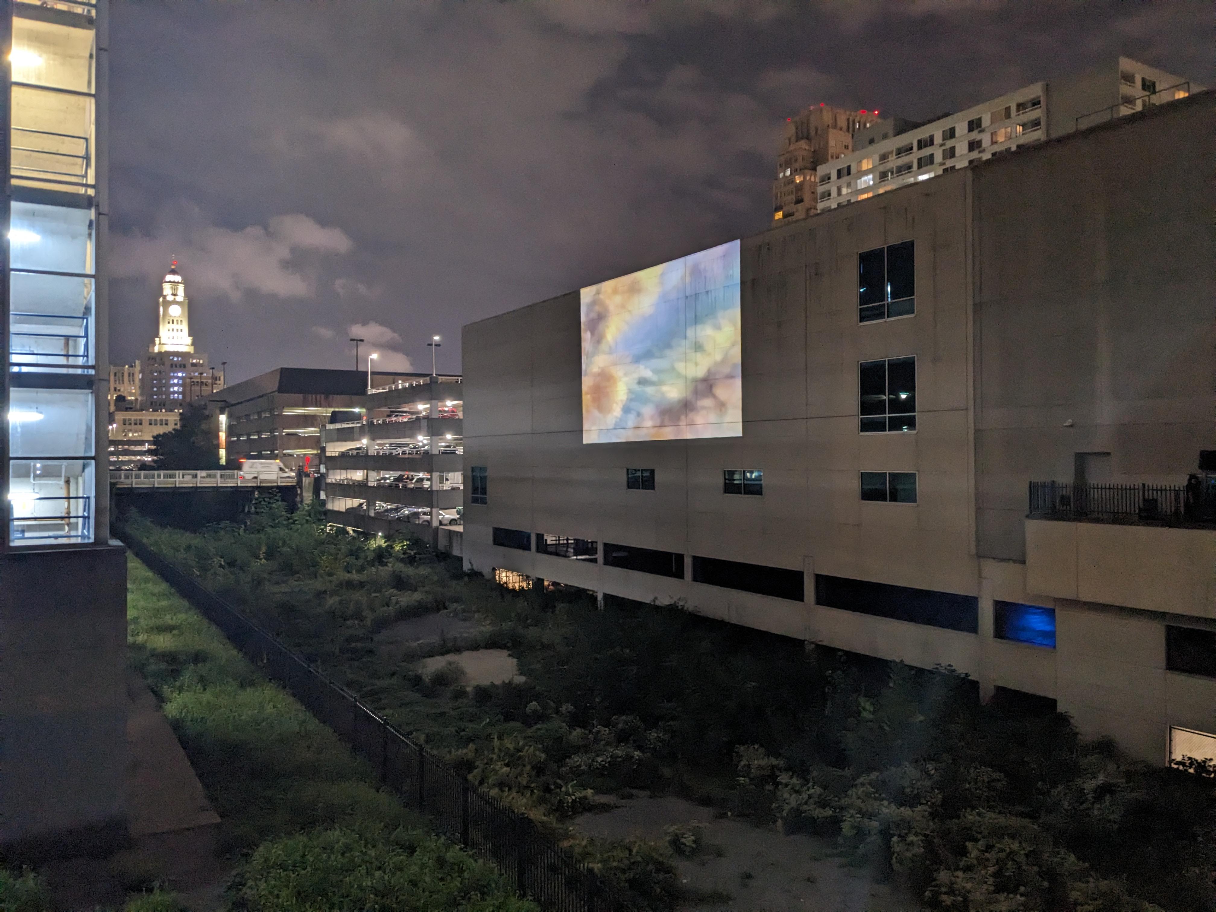 Photo courtesy of eos. Night time photo of the overgrown section surrounded by building with colorful projection on the wall.