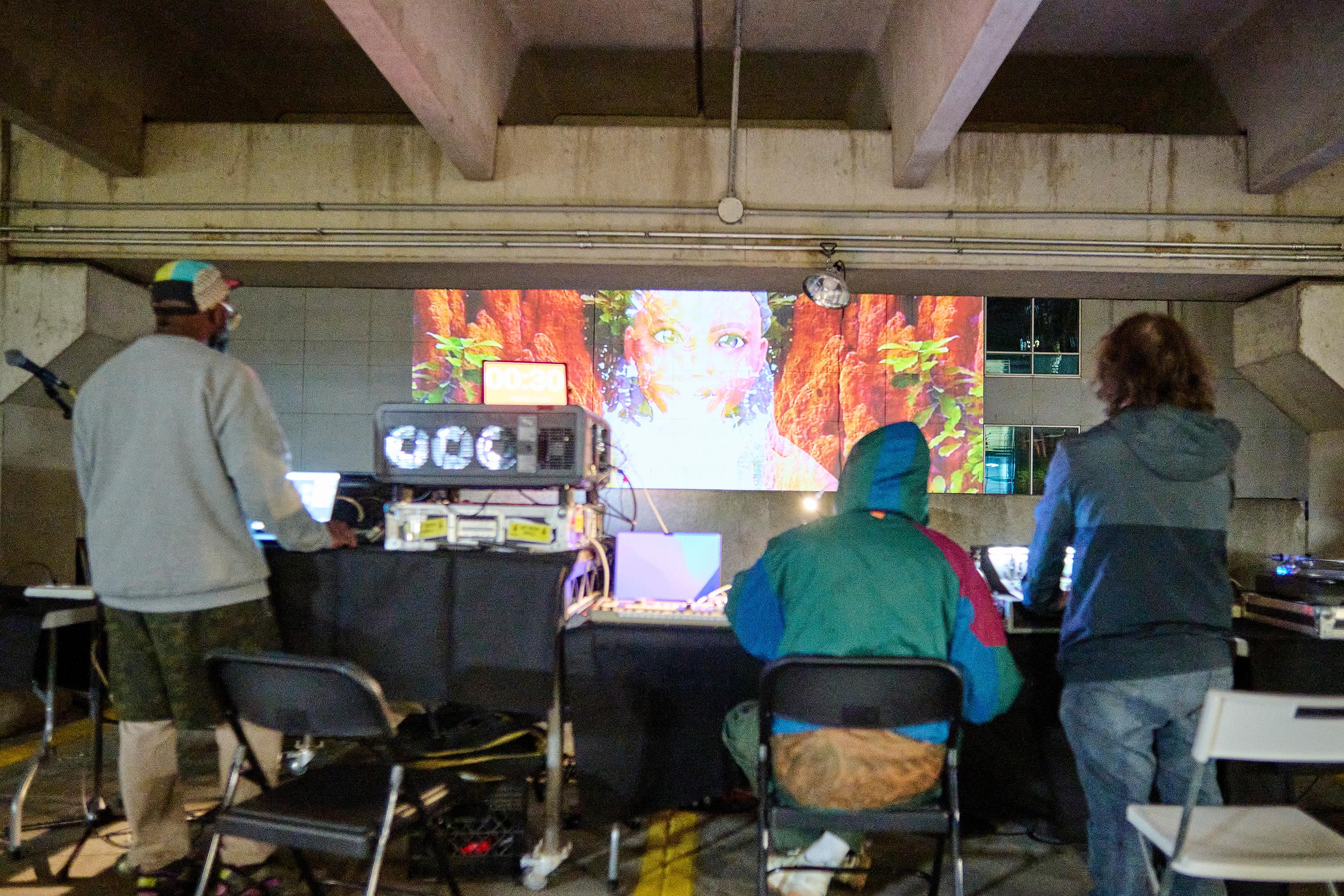 Photo by Albert Yee. Three people work at a table with computers and audio equipment DJing and coordinating a public video art installation projected onto the face of a building in front of them.