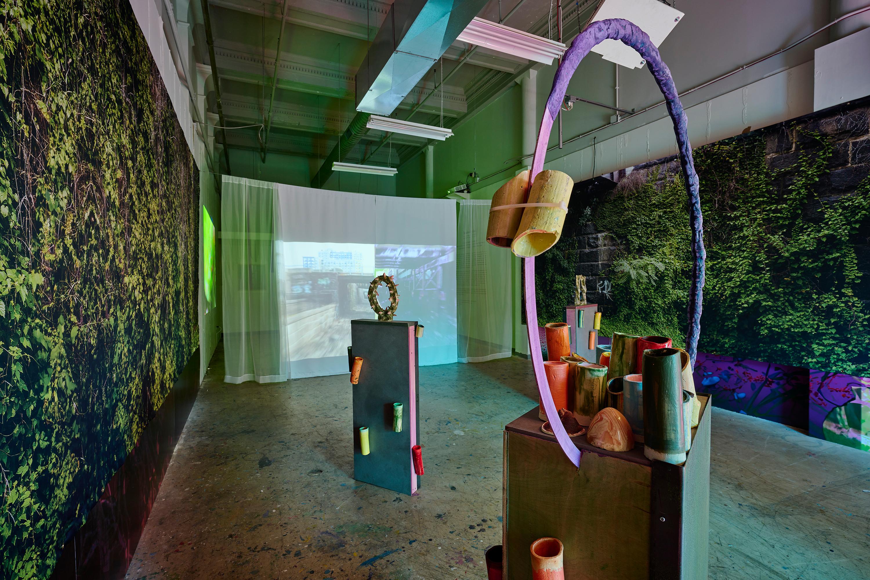 Photo courtesy of Albert Yee. Picture of the invasive species gallery, showing the walls with the vine backdrop, the 2 sculptures in the middle of the room, and projector with picture of the cut.