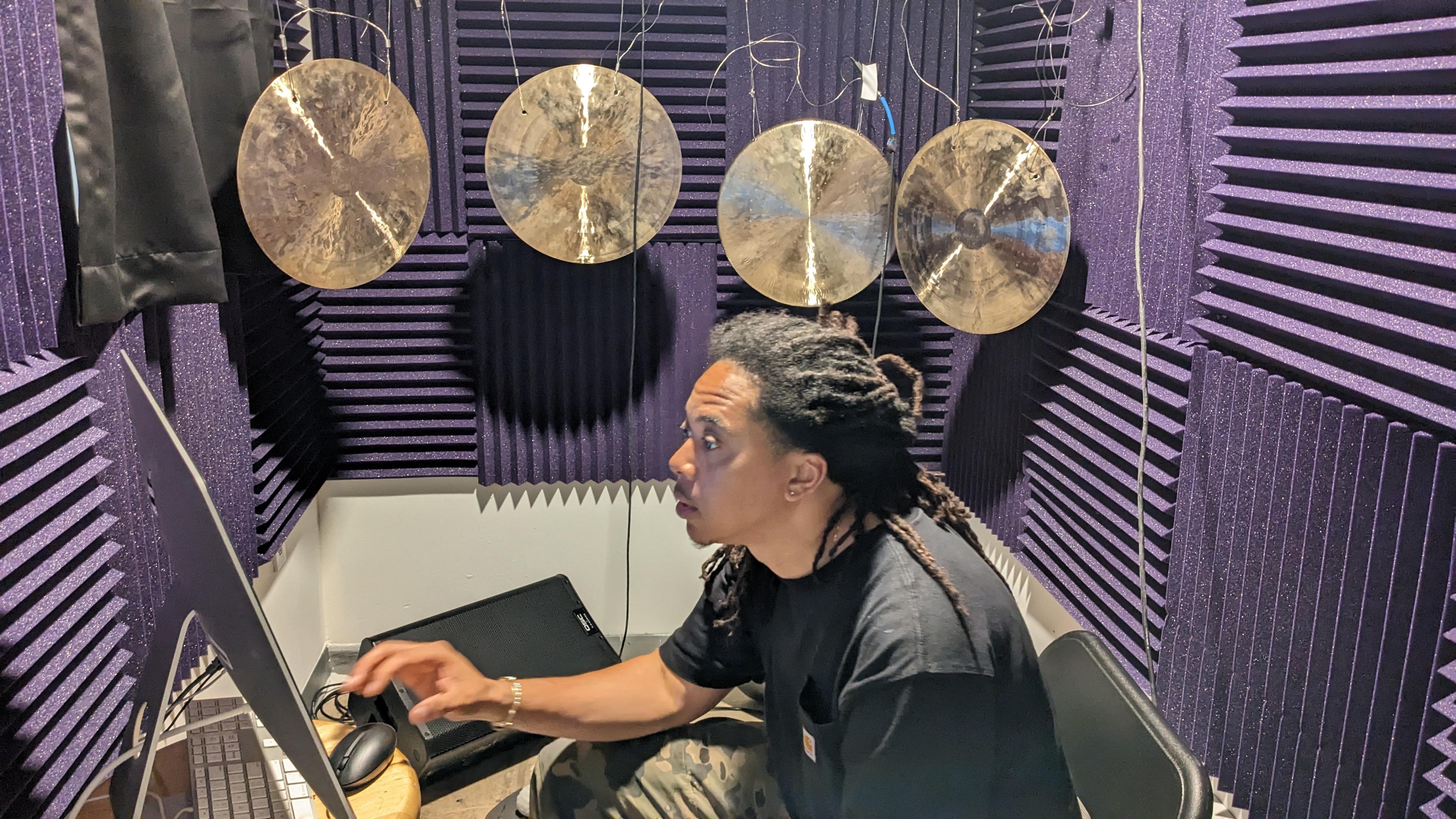 Photo by eo Studios. A person sits at a computer in a sound booth sound engineering with gongs hanging in the back.