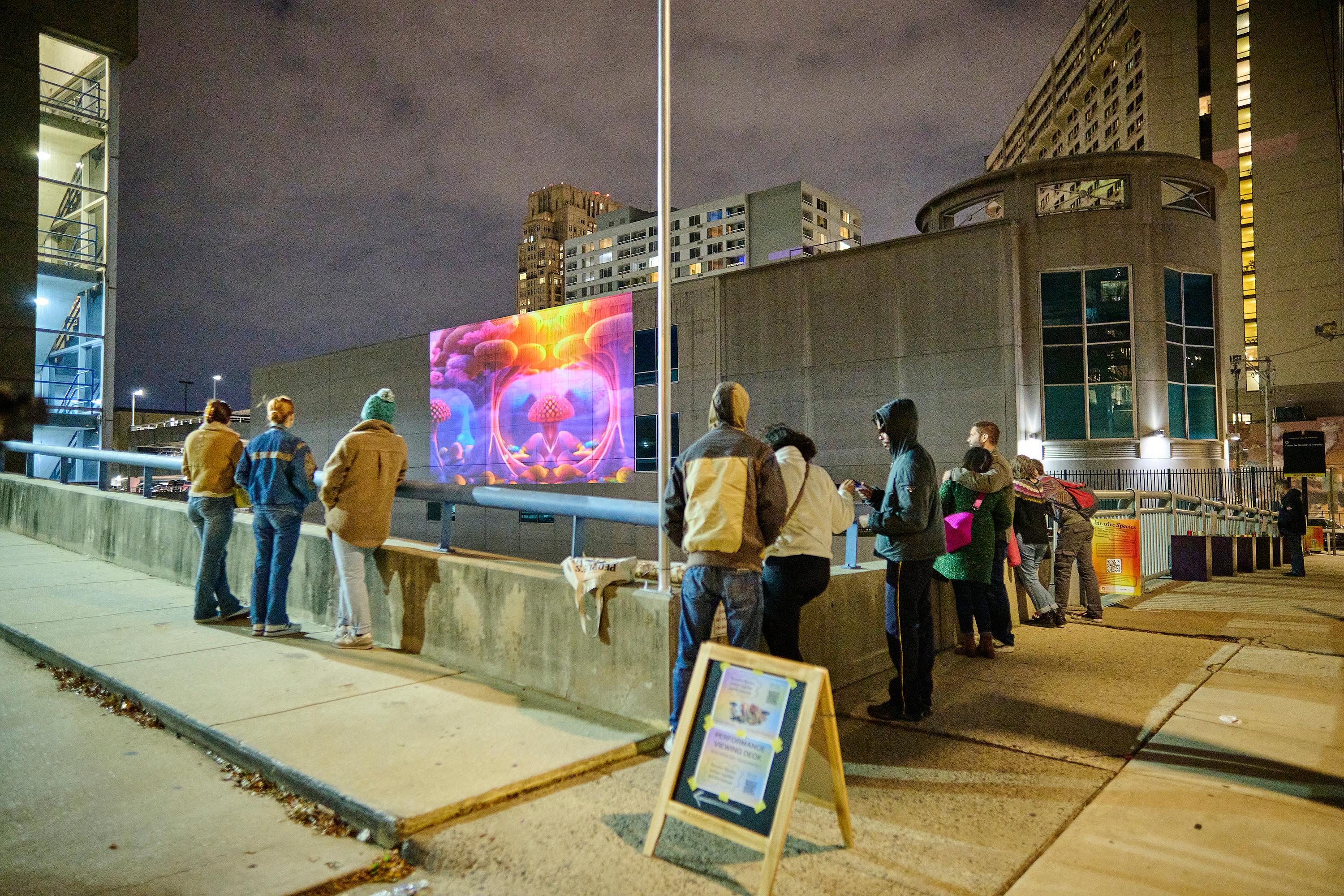 Photo courtesy of Albert Yee. Night time picture of people looking at a projection of the exhibition against the building wall.