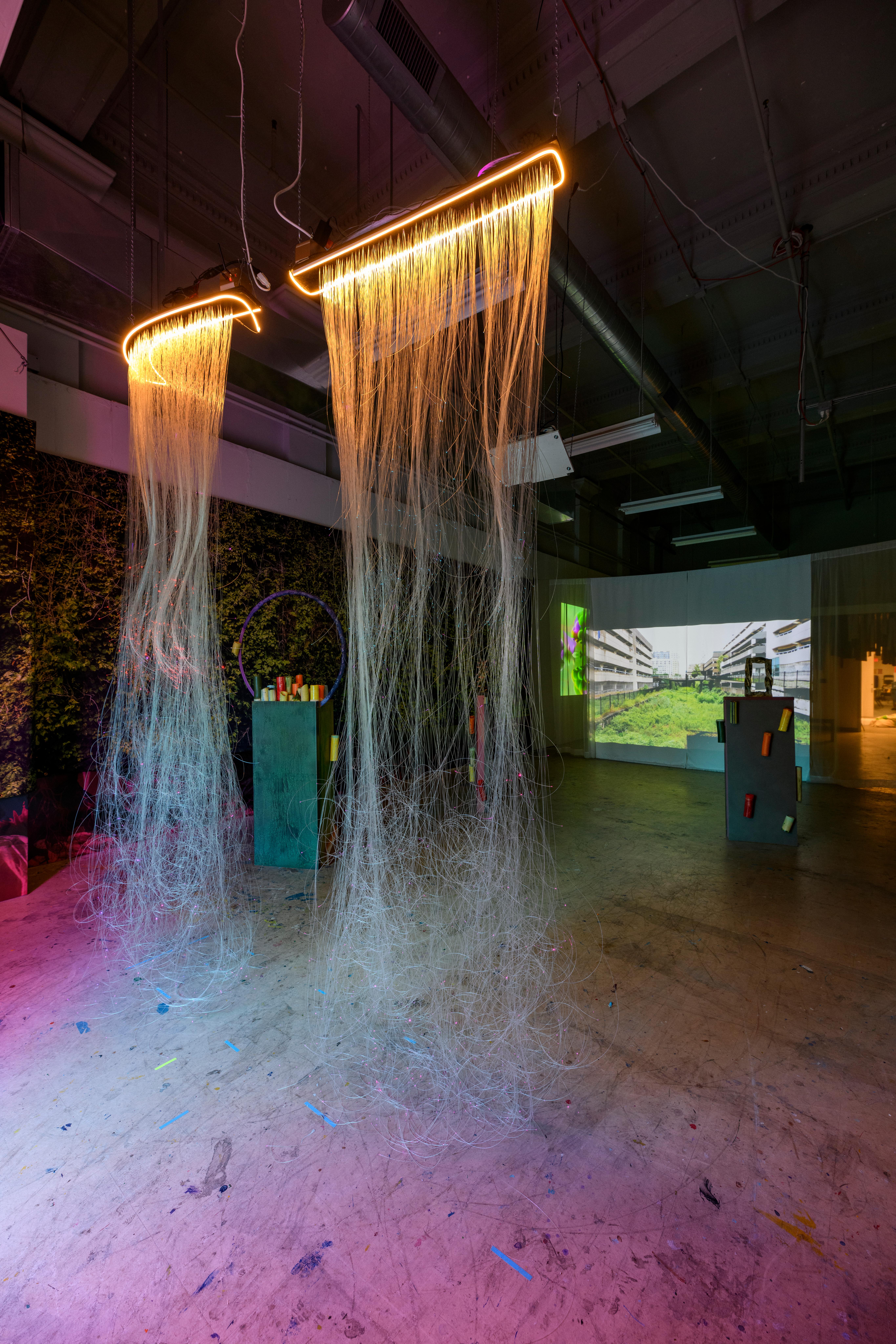 Photo by Constance Mensh. A view of a gallery with multiple sculptural installations and video projection in the back. A piece with plastic strings hang in the foreground lit with LEDs at the top.