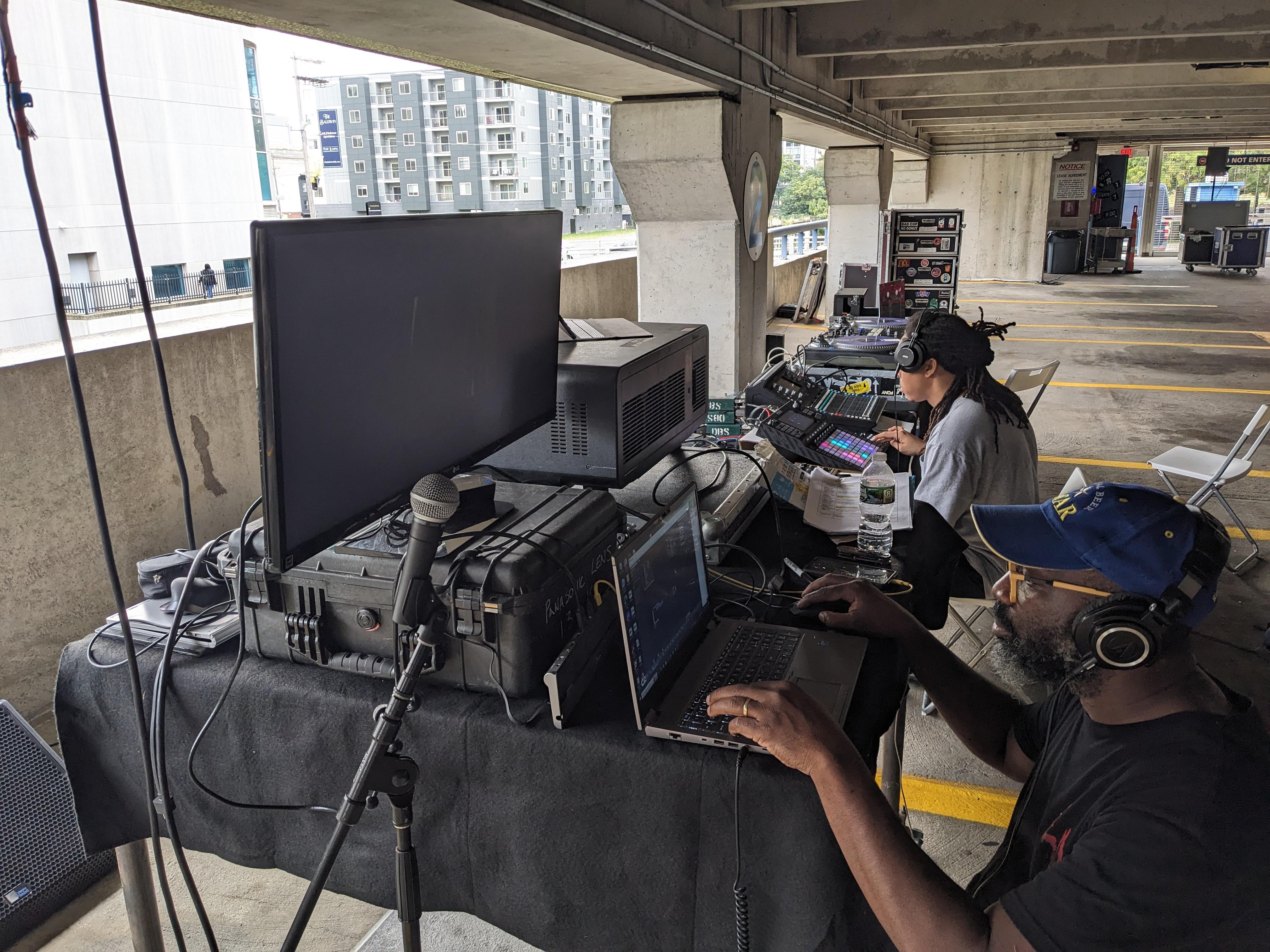 Photo by eo Studios. Two people work at a table covered in computers and audio equipment, inside a parking garage.