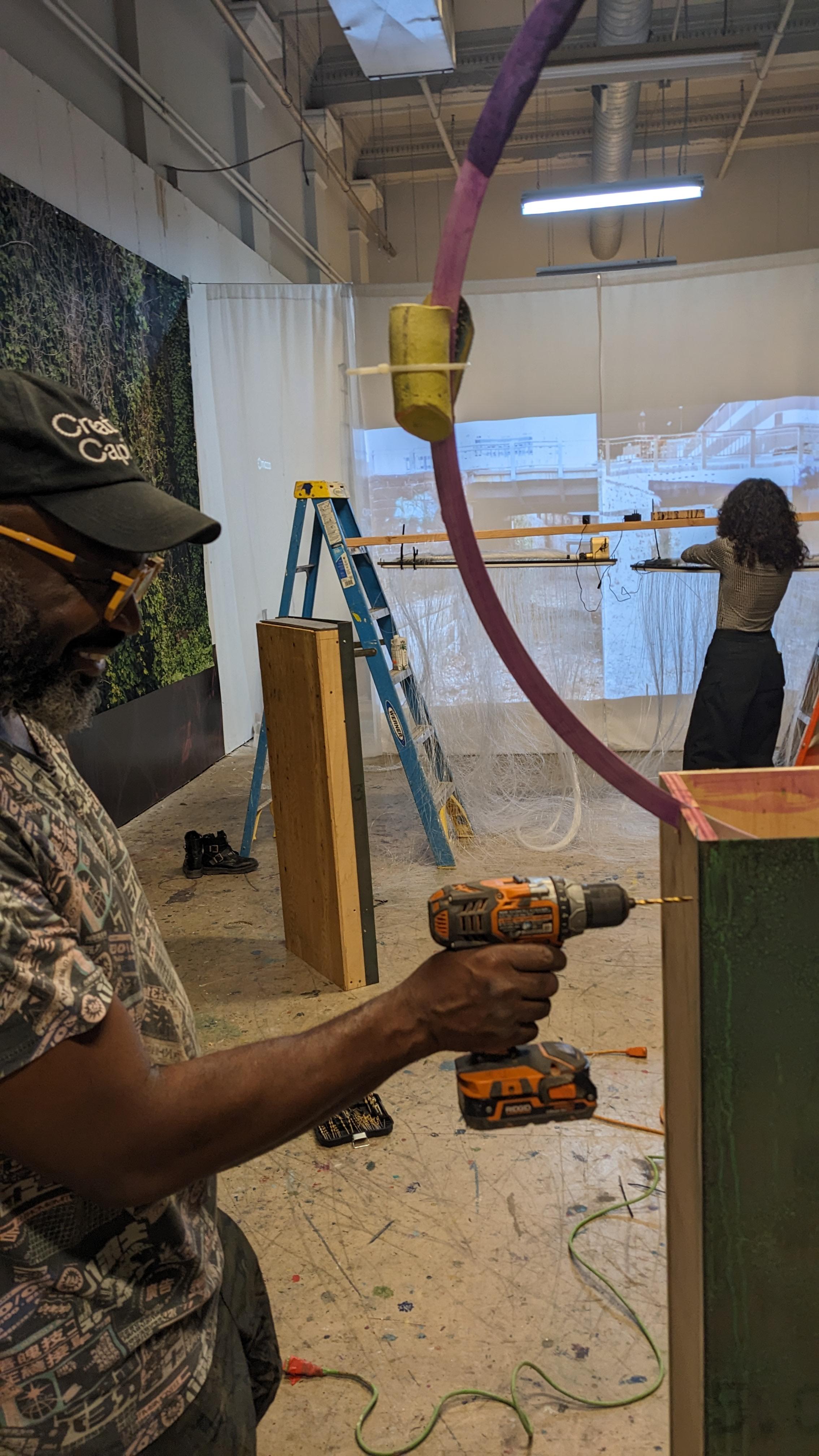 Photo by eo Studios. Photo of a gallery space during the installation of an exhibit. Multiple people work on the install. A person is drilling in the foreground.